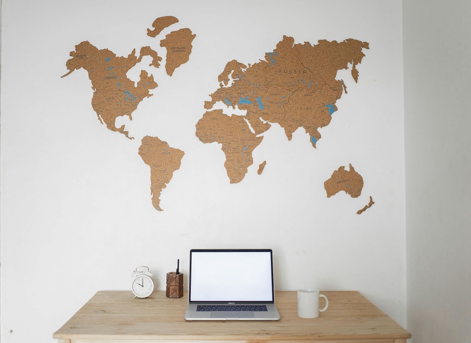 A laptop with a blank white screen sits on a desk in front of a wooden world map on a white wall.
