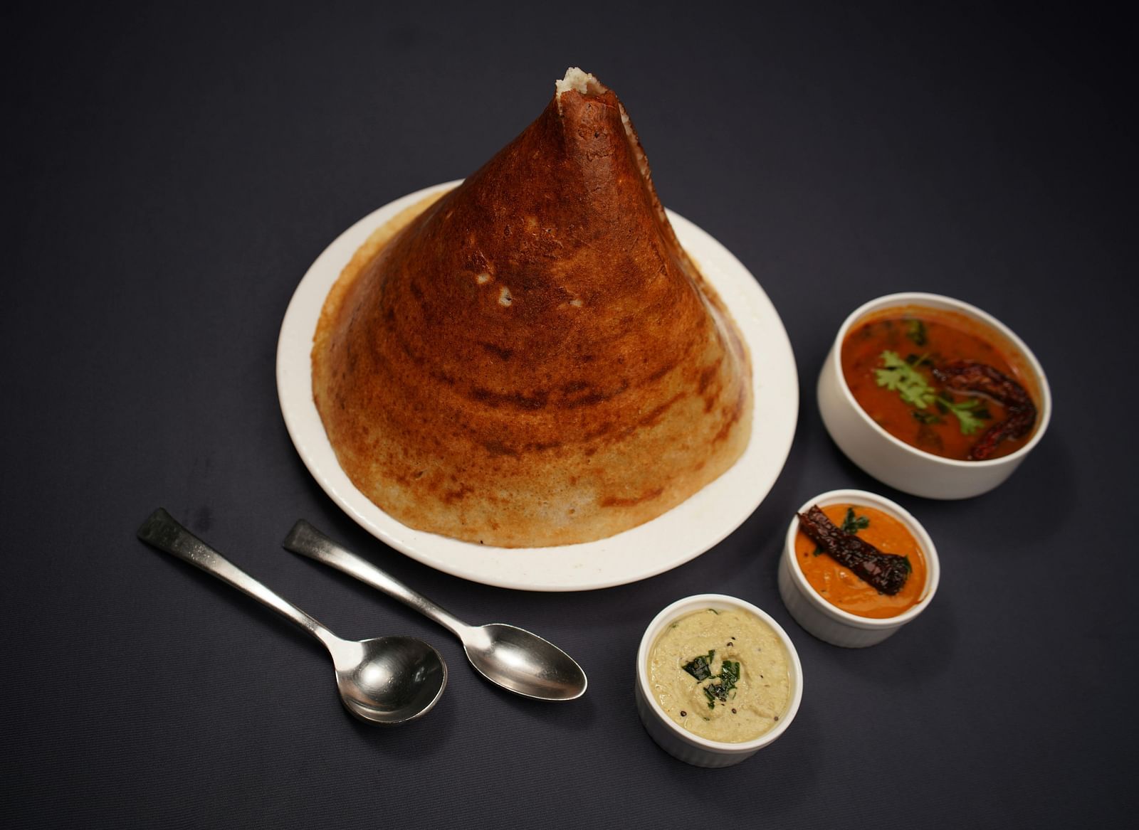 A golden dosa on a white plate, accompanied by three small bowls of colourful chutneys and sambar, with two spoons resting beside it.