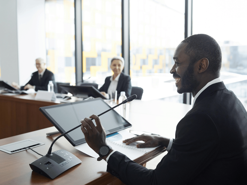 A professional man in a suit speaking into a microphone at a conference table, smiling, with two colleagues seated across from him and large windows in the background.