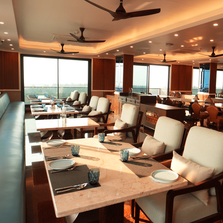 Indoor restaurant seating with cushioned chairs, tables set for dining, and ceiling fans overhead at MAYFAIR Bay Resort, Paradeep.