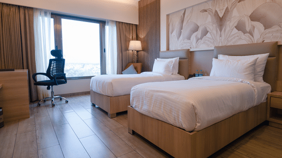 A bright hotel room featuring twin beds, a wall-mounted piece of art above the beds, and a large window offering outdoor views | Pride Elite Chhatrapati Sambhajinagar