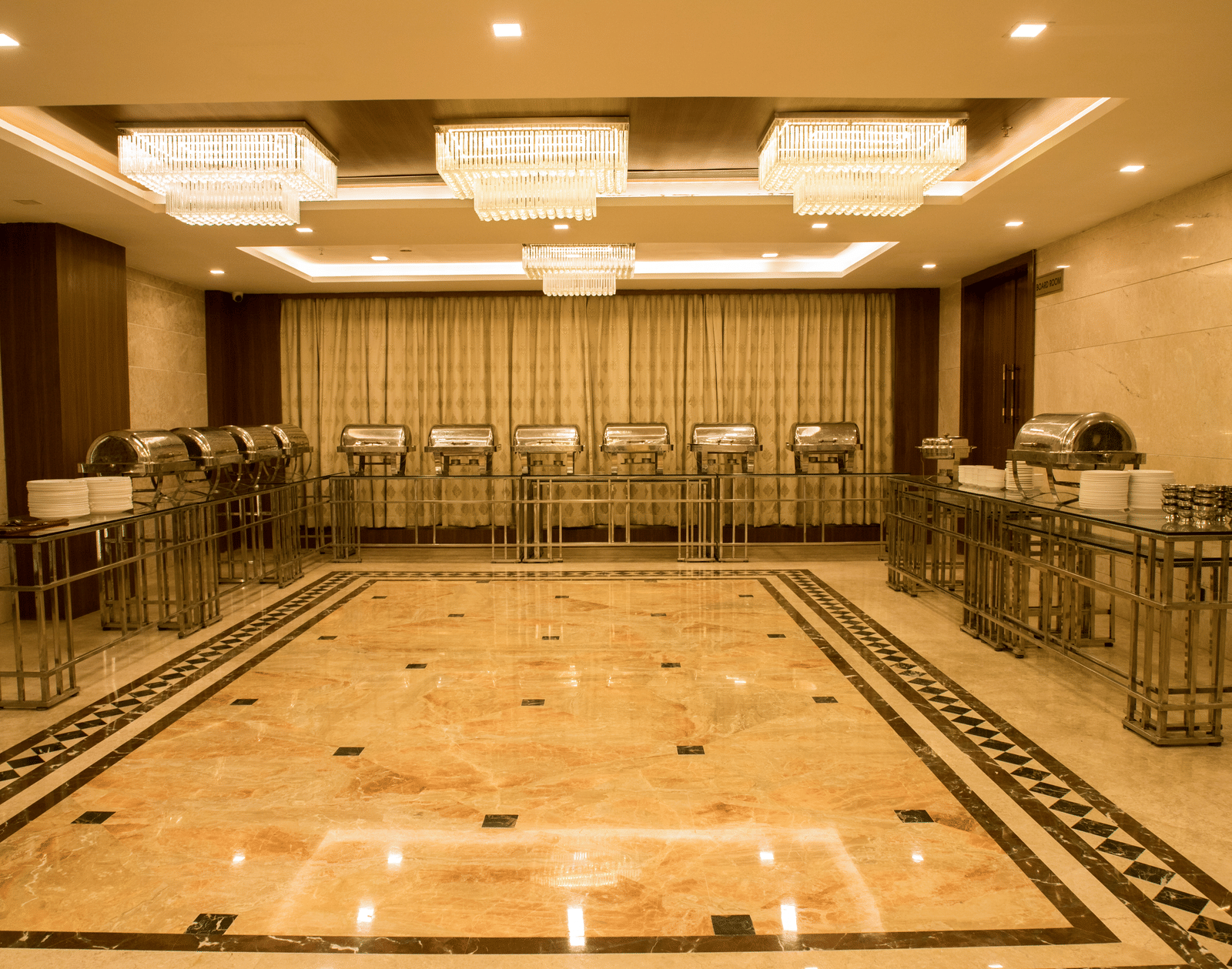 A brightly lit banquet hall with several chafing dishes lined at the far end of the hall at Benzz Park, Vellore.