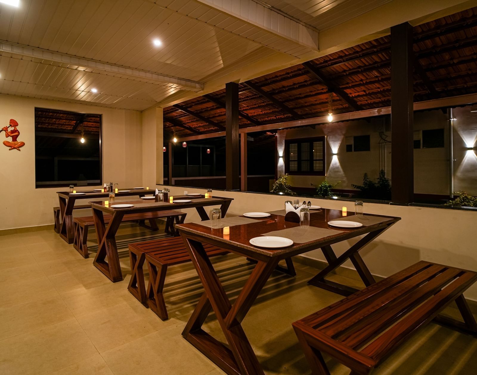 An evening shot of the Mane Cafe at Bhuthanakadu Retreat, showing a well-lit, open-air dining space with rustic wooden tables and bench seating.