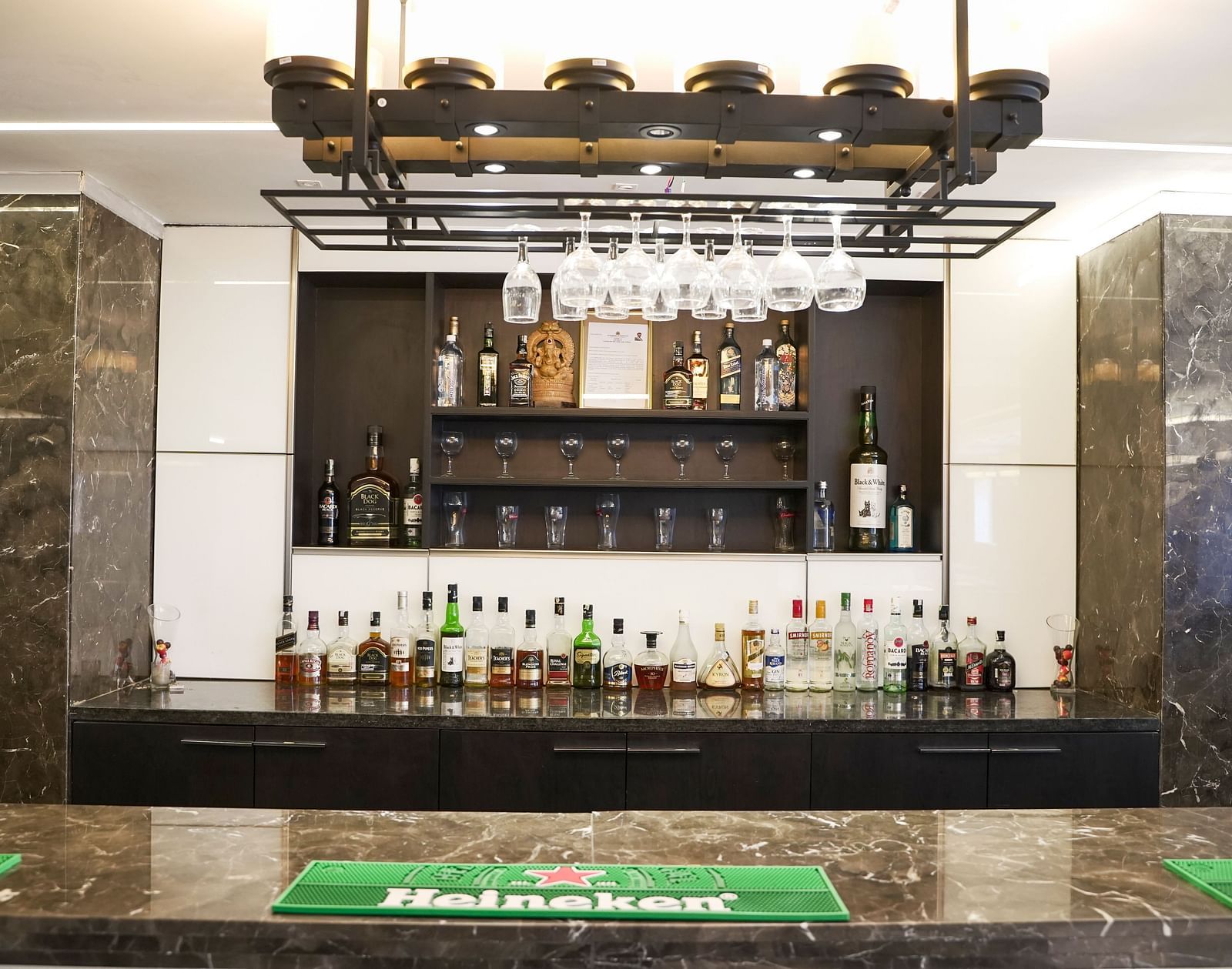 A complete view of a fully stocked bar at BNGV Grandeur Hotel and Banquets, with an impressive display of bottles, a green-lit counter, and an overhead rack for glasses.