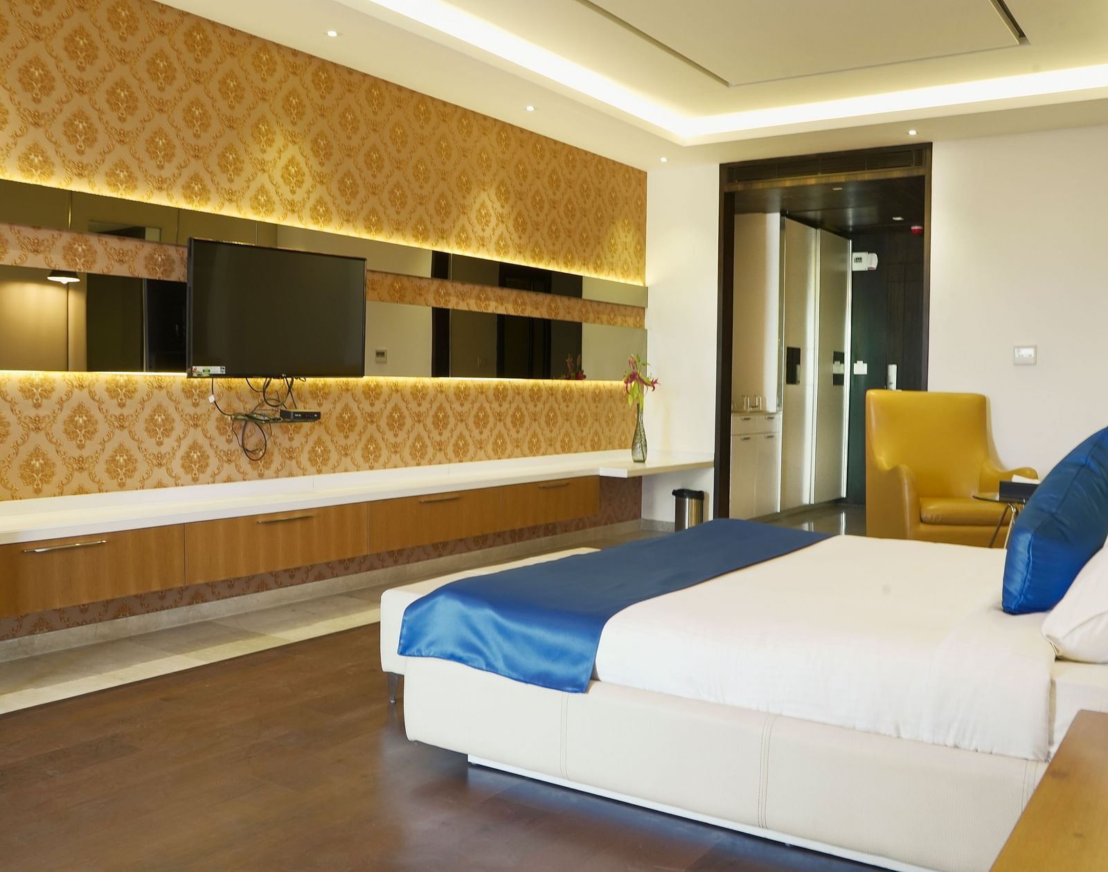 A well-appointed hotel room at BNGV Grandeur Hotel and Banquets, featuring a bed with blue and white linens, a patterned wall, and a dresser with a mirror.