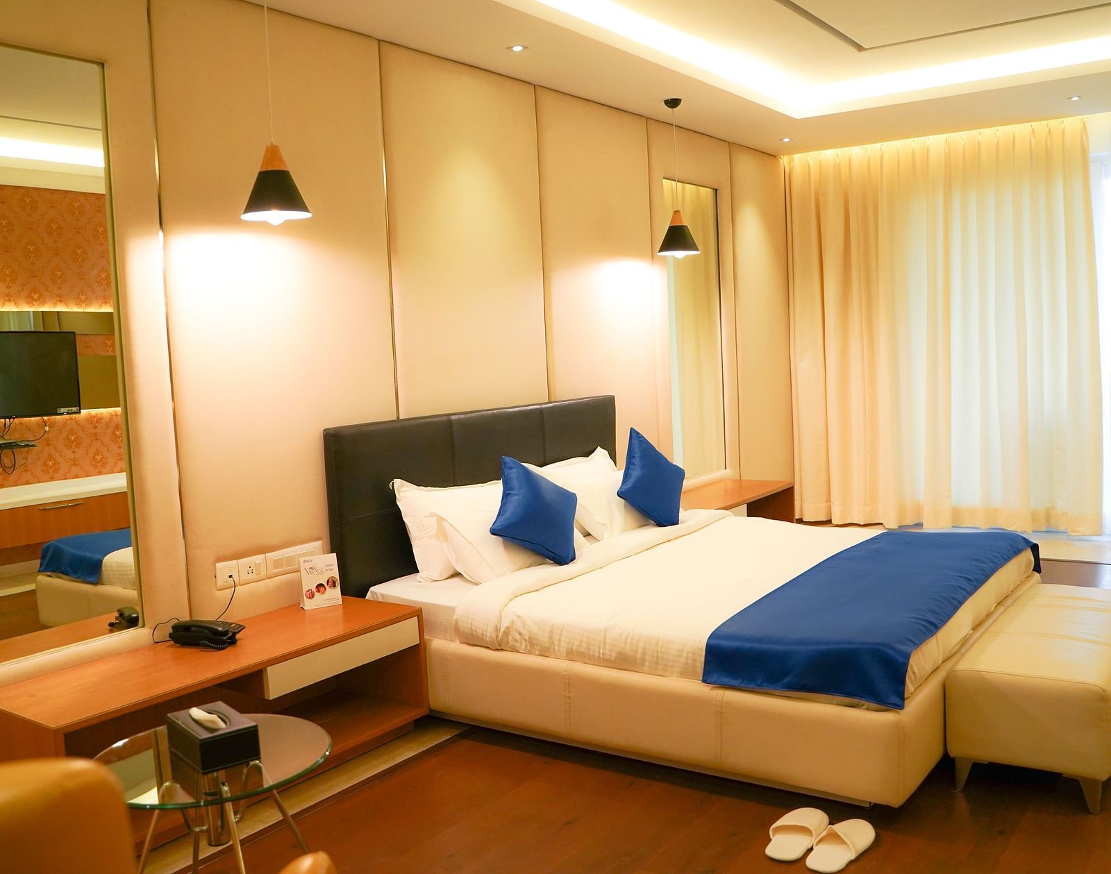 A warm-toned hotel room at BNGV Grandeur Hotel and Banquets, with a bed featuring blue and white linens, and soft lighting creating a cosy ambience.