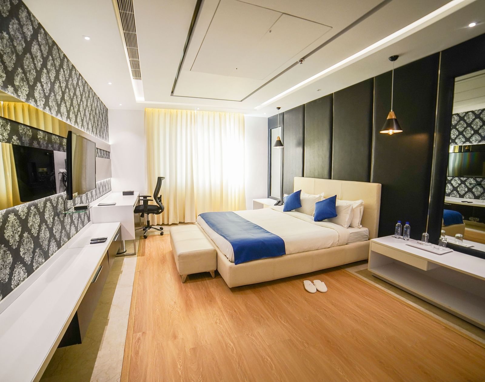 A modern and elegantly designed hotel room at BNGV Grandeur Hotel and Banquets, featuring a comfortable bed with a blue runner, a unique patterned accent wall, and contemporary furnishings.