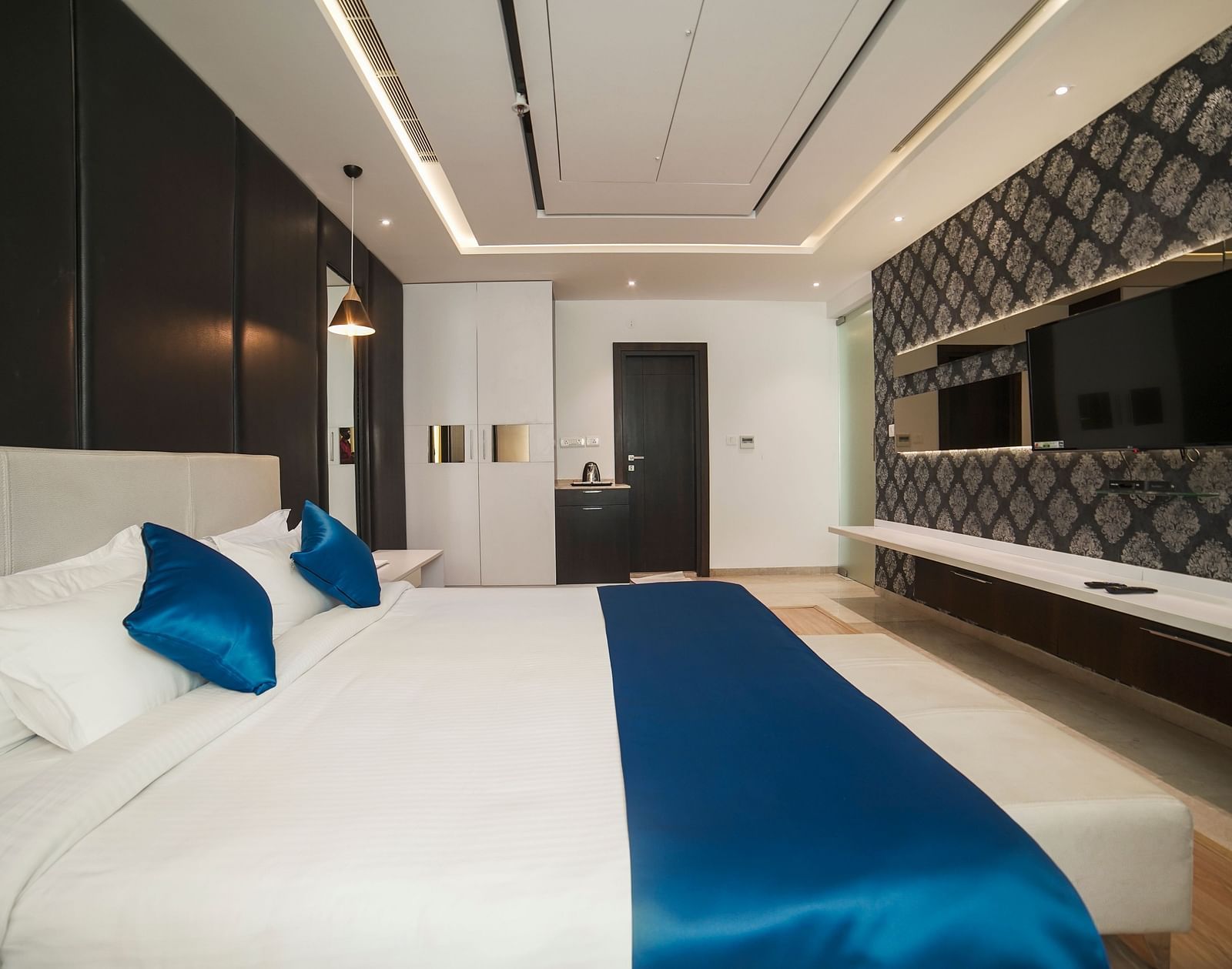A modern hotel room at BNGV Grandeur Hotel and Banquets, featuring a comfortable bed with a blue bed runner, clean white linens, and sleek furnishings.