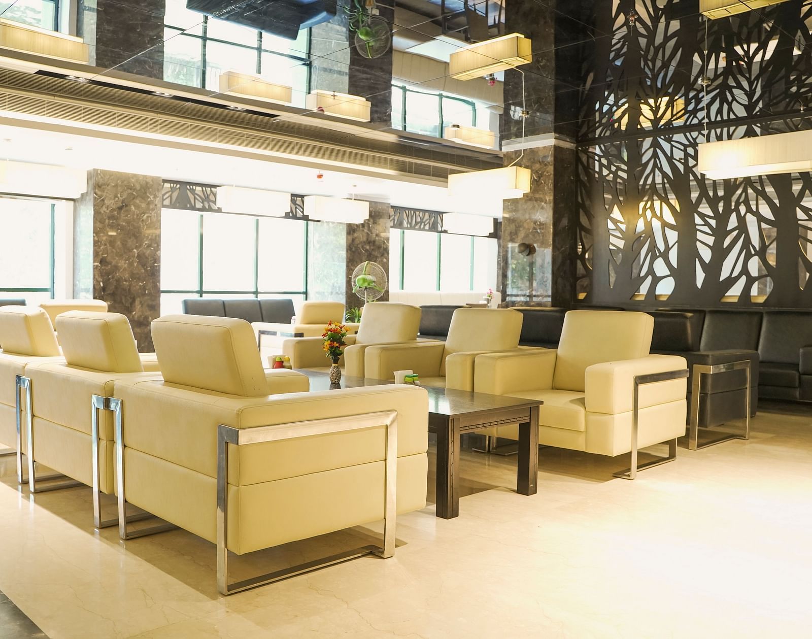 A broad view of a luxurious lounge at BNGV Grandeur Hotel and Banquets, featuring contemporary light-coloured seating, dark decorative wall panels, and natural light from large windows.