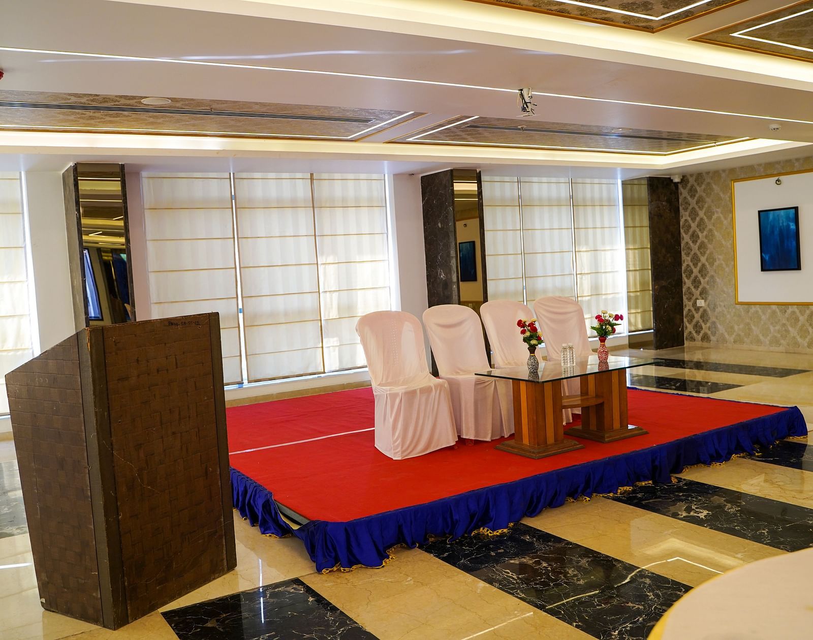 A banquet hall at BNGV Grandeur Hotel and Banquets, set up with a raised red stage, a podium, and chairs, ready for an event.
