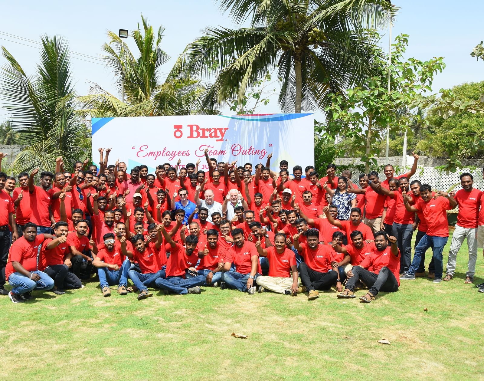 A group of people wearing red t-shirts posing on an outdoor lawn with a banner featuring green trees at Grande Bay Resort & Spa.