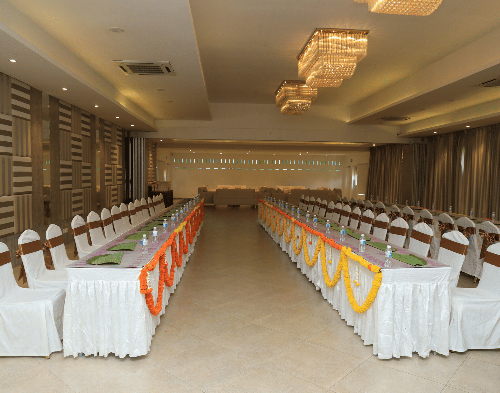 A long indoor banquet hall with multiple long tables arranged for an event, featuring white chairs with sashes and colourful table runners - Grande Bay Resort & Spa, Mamallapuram