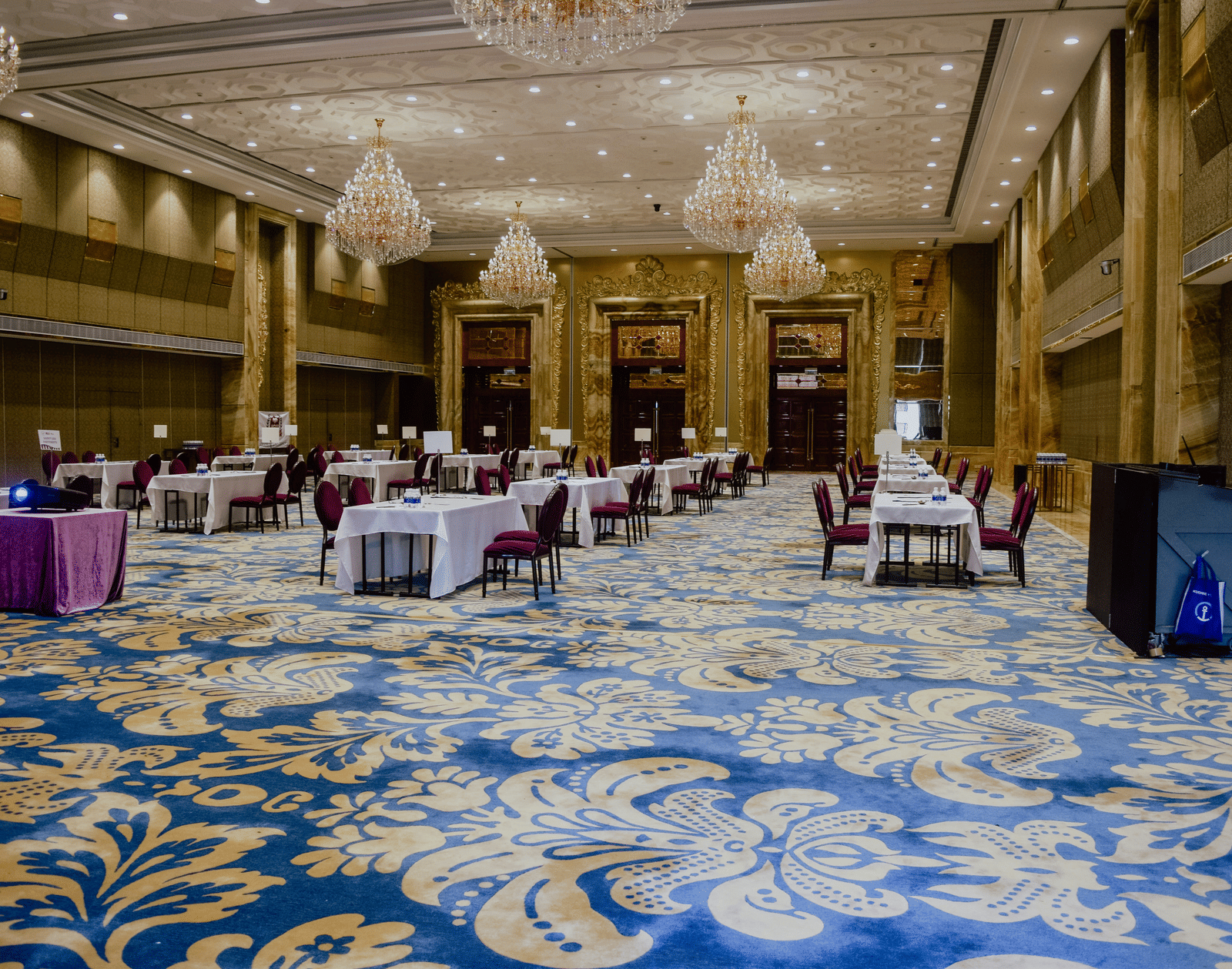 A large hall with patterned blue carpet, multiple chandeliers, and sets of tables and chairs