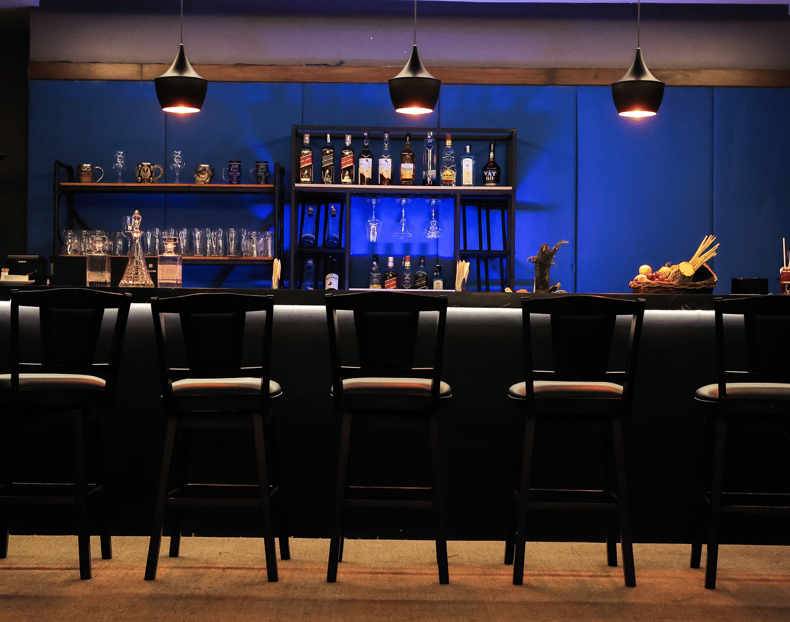 Wide view of the dark, sleek bar with a blue accent wall, shelving full of bottles, and black leather bar stools at Hotel Hukam's Lalit Mahal.