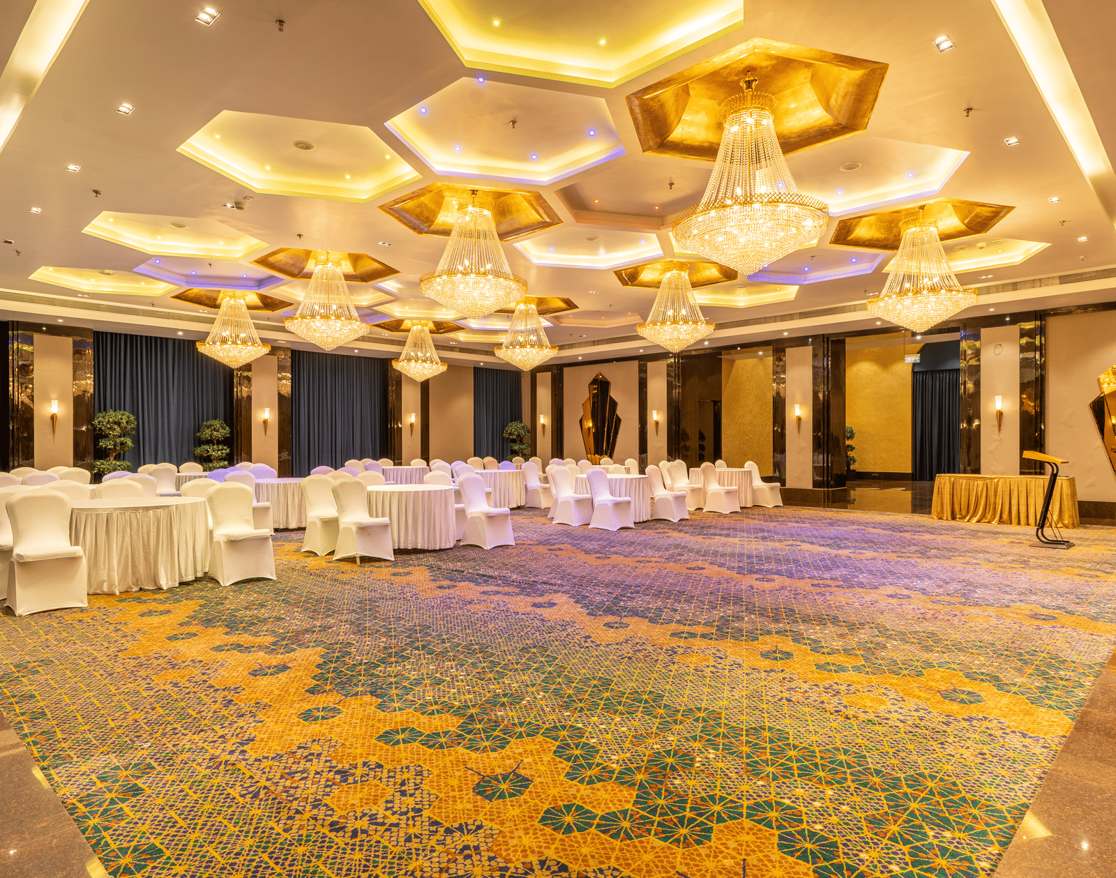 Wide view of a long banquet table covered in white cloth, set up under ornate gold and white hexagonal chandeliers at Hotel Hukam's Lalit Mahal.