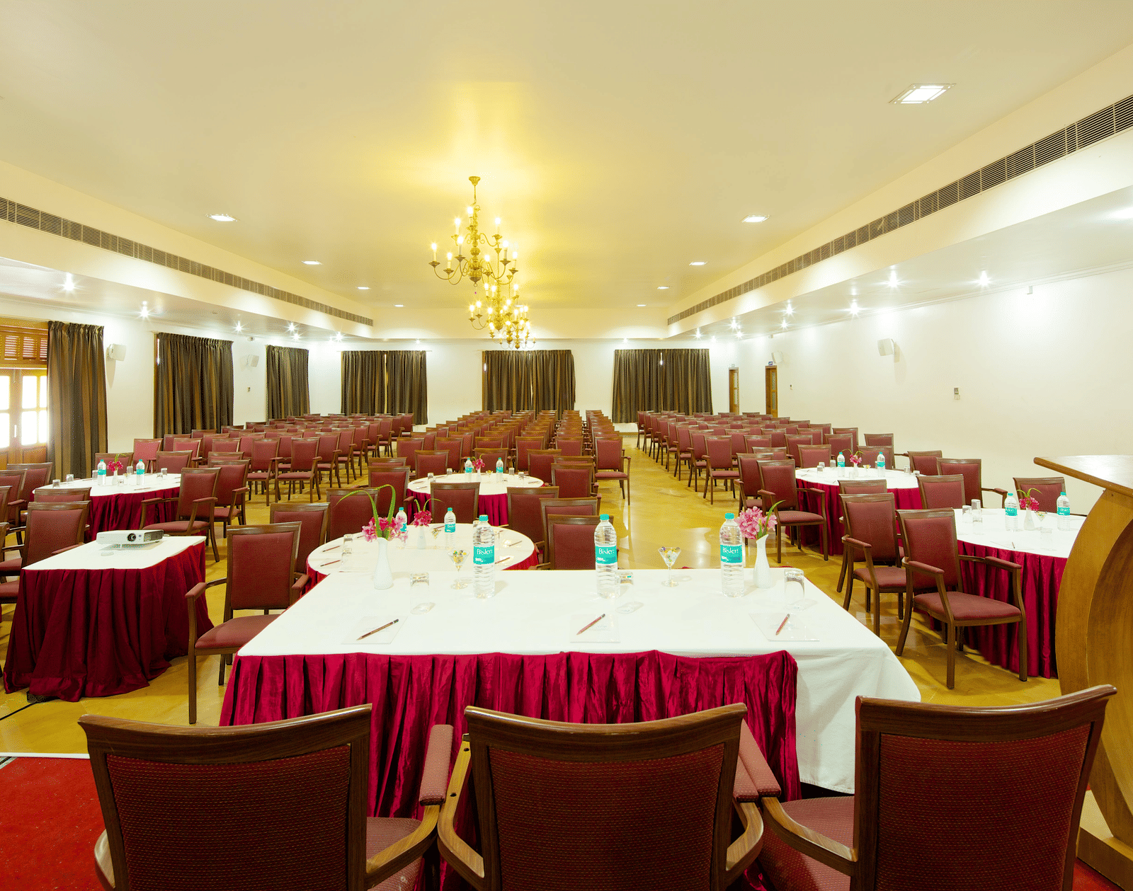 A large meeting room arranged with multiple tables covered in white tablecloths with red valances and surrounded by chairs | Le Pondy