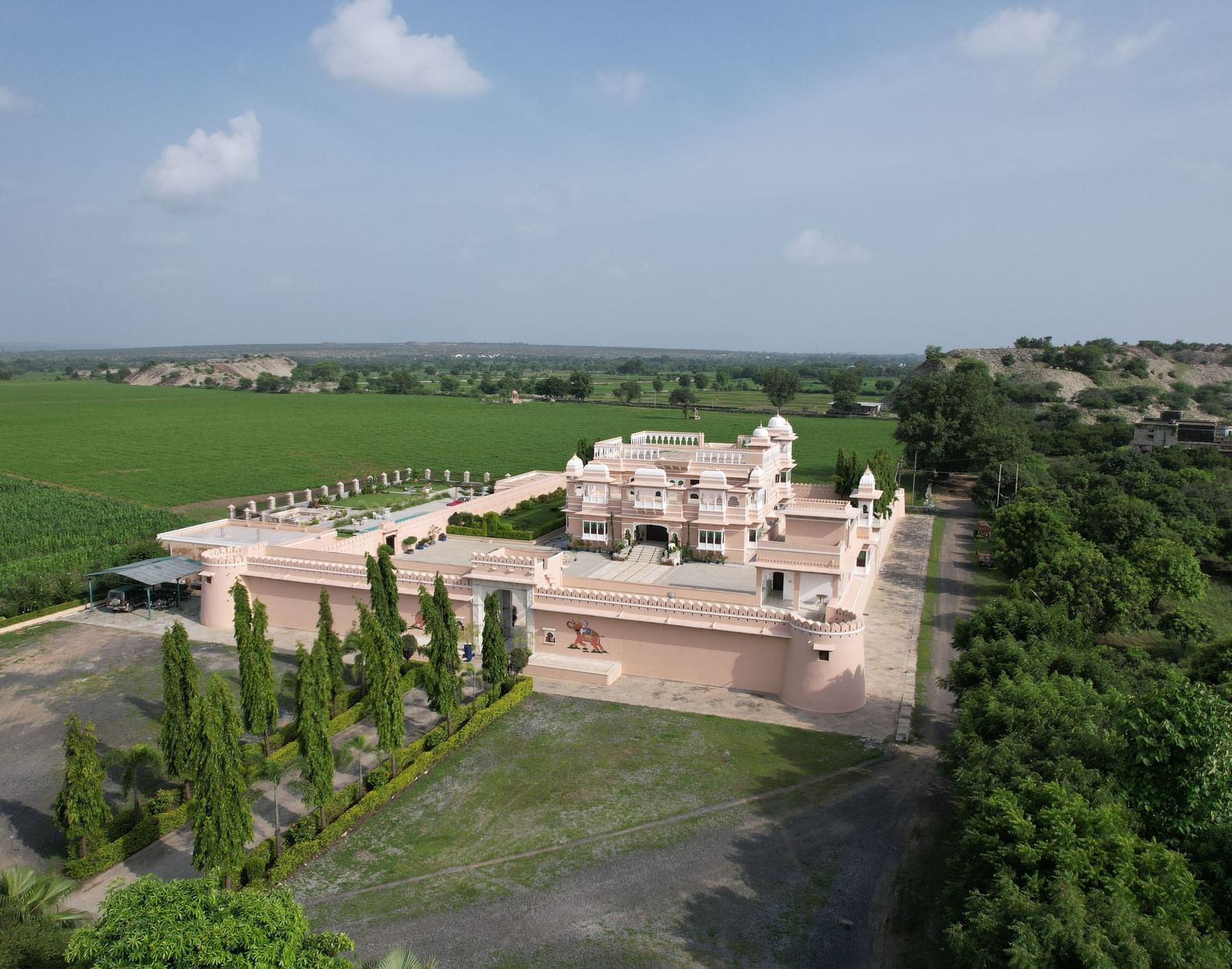An aerial view of a large, light-colored building complex surrounded by green trees and fields at Mahendra Niwas. A paved drive and a landscaped garden are visible in the foreground.