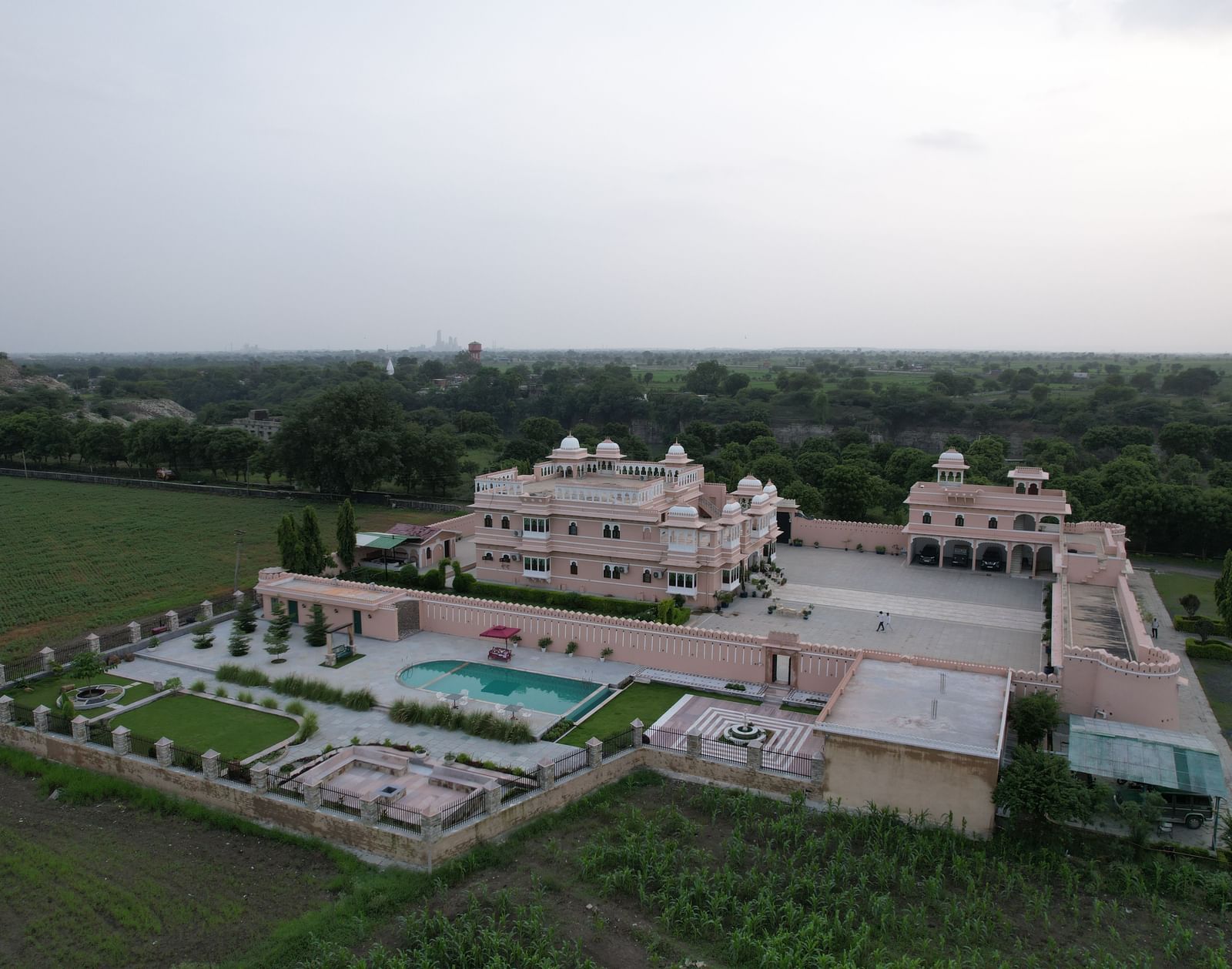 A high-angle aerial view of Mahendra Niwas, presenting the entire property with its intricate garden design, swimming pool, and surrounding landscape, all under a clear sky.