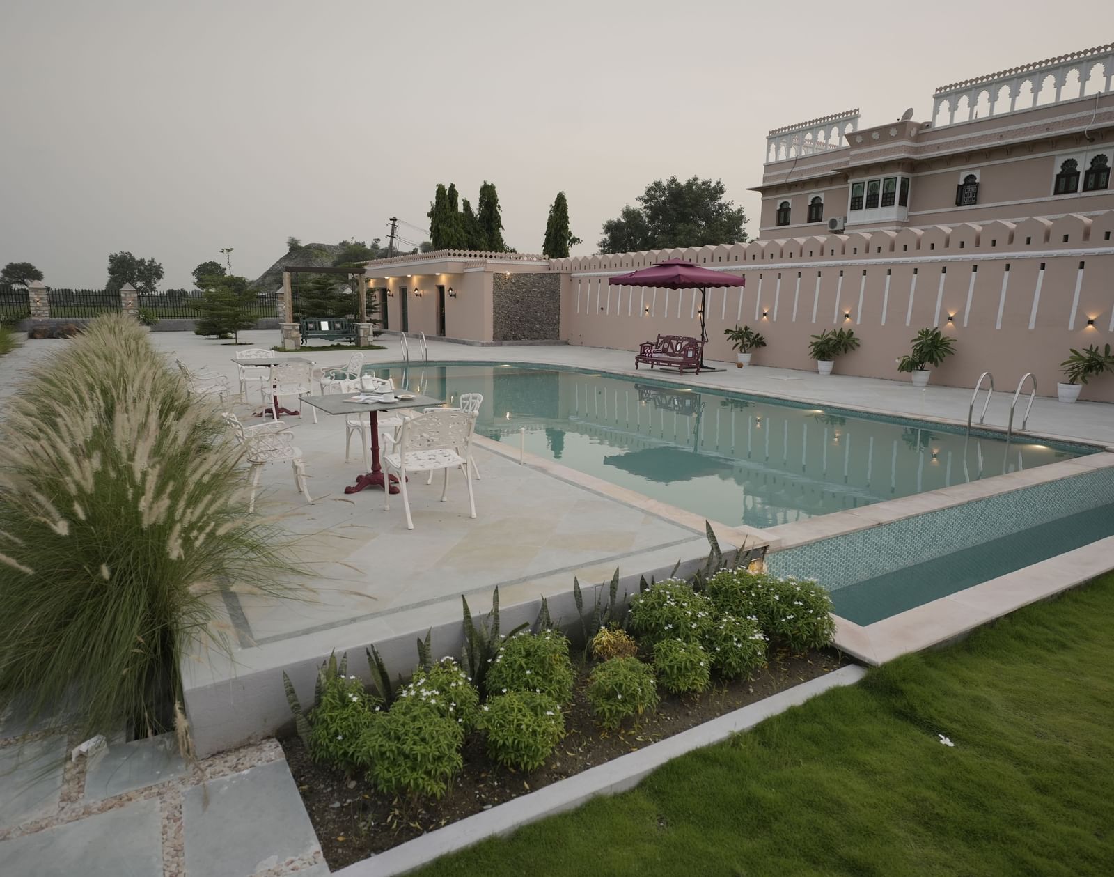 A wide, eye-level shot of a pool surrounded by a large grassy area at Mahendra Niwas. The water is a bright turquoise, and a building with a terrace is visible in the background.