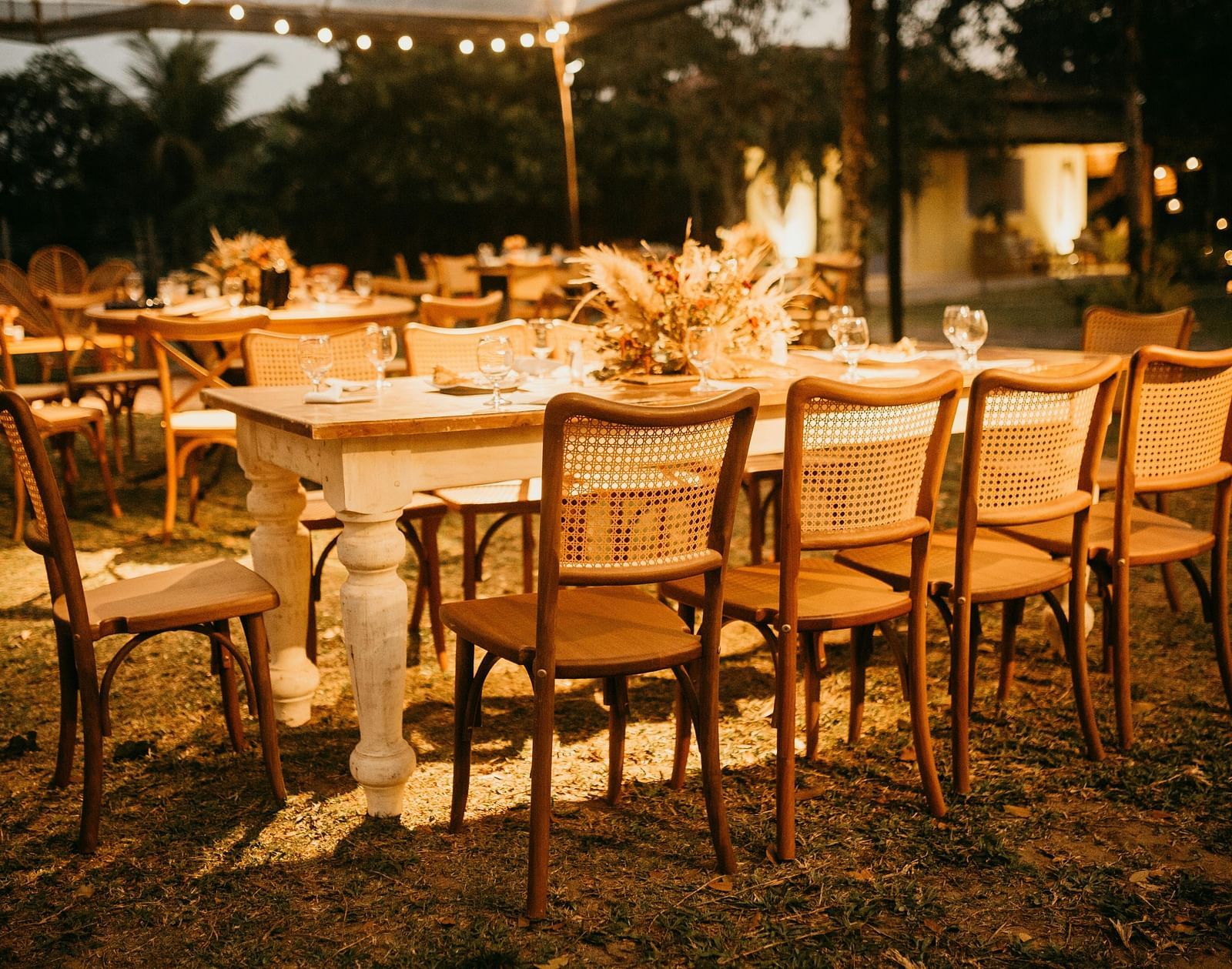 An outdoor setup featuring long tables with wooden chairs, ambient lighting, and a natural setting.