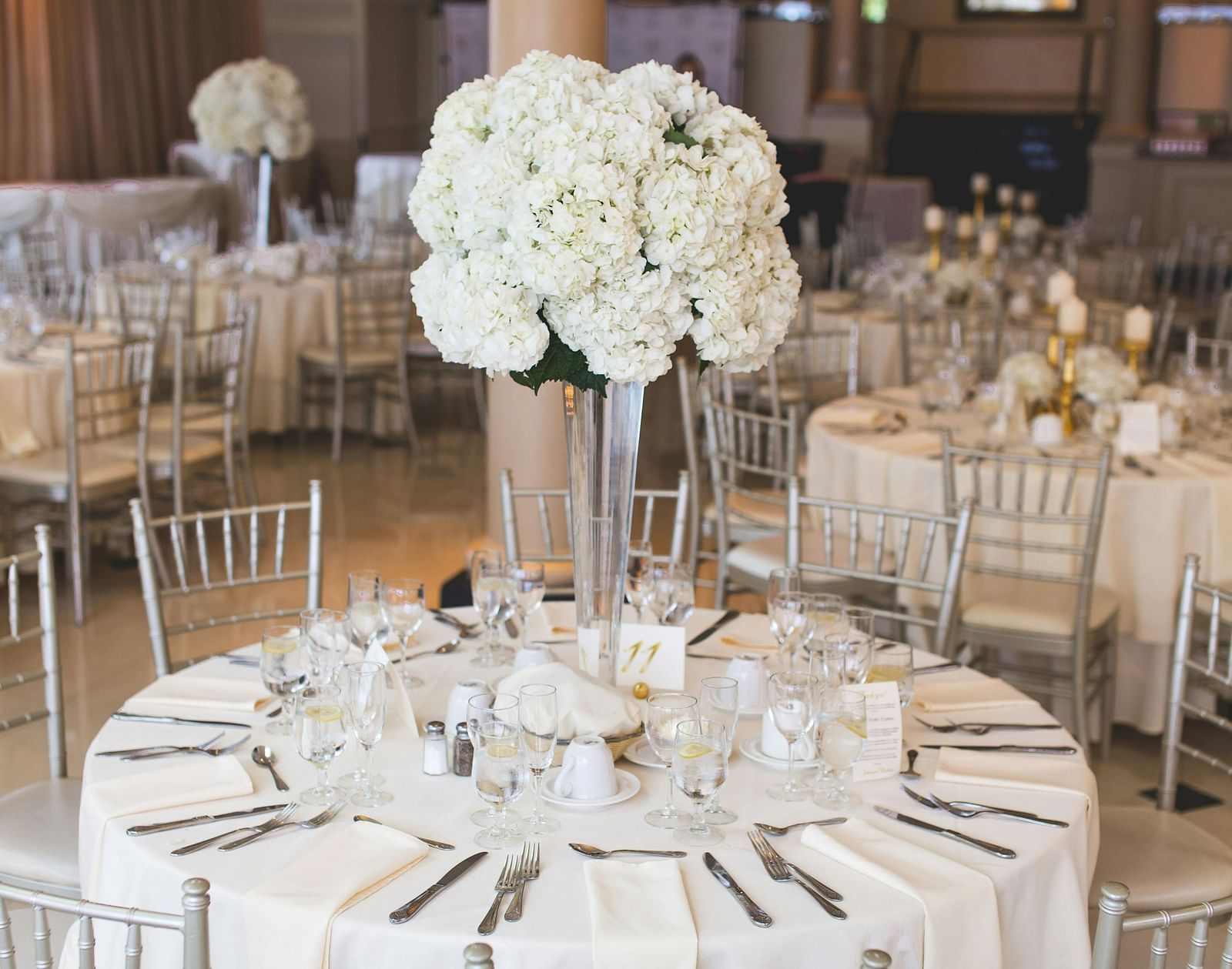 An indoor reception displaying round tables with white linens, floral centrepieces, and coordinated place settings.