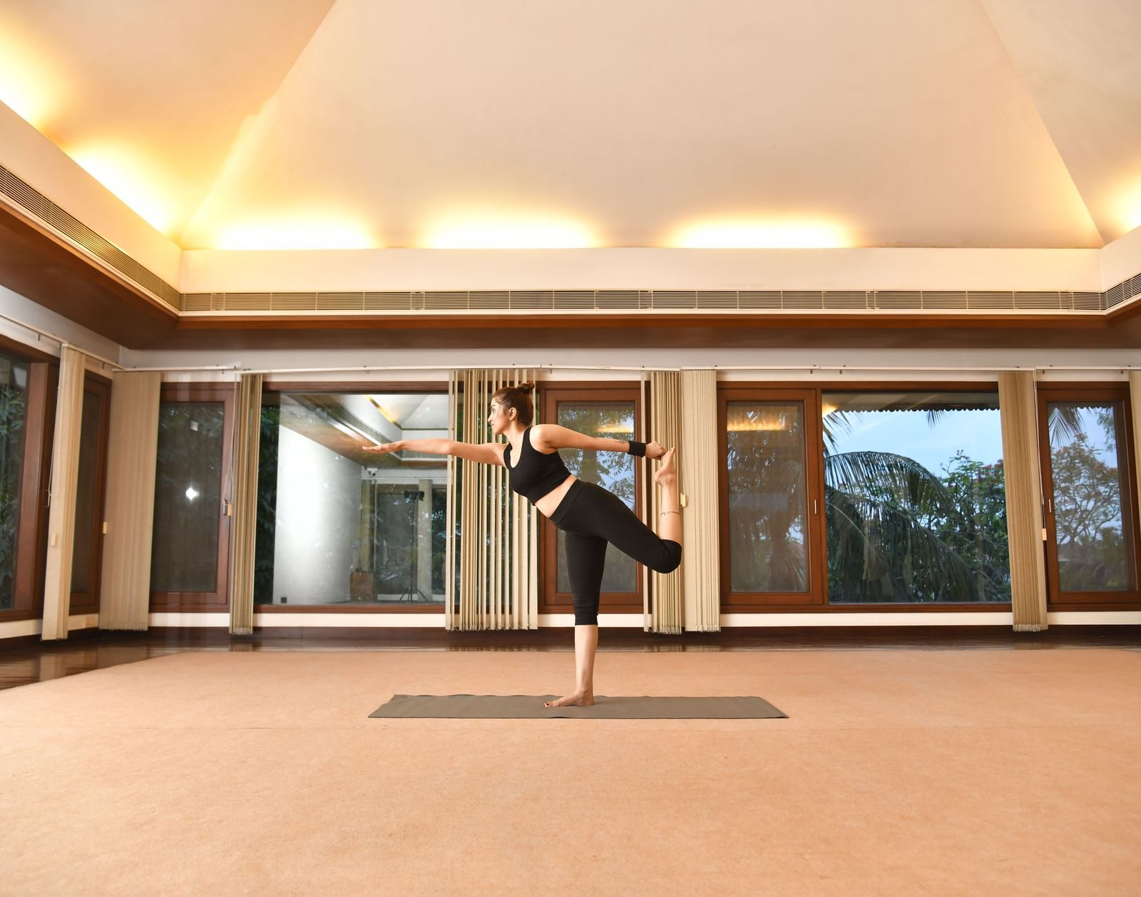 A woman in black activewear practices a balancing yoga pose on one leg in a spacious room with wooden floors, large windows, and mirrored walls