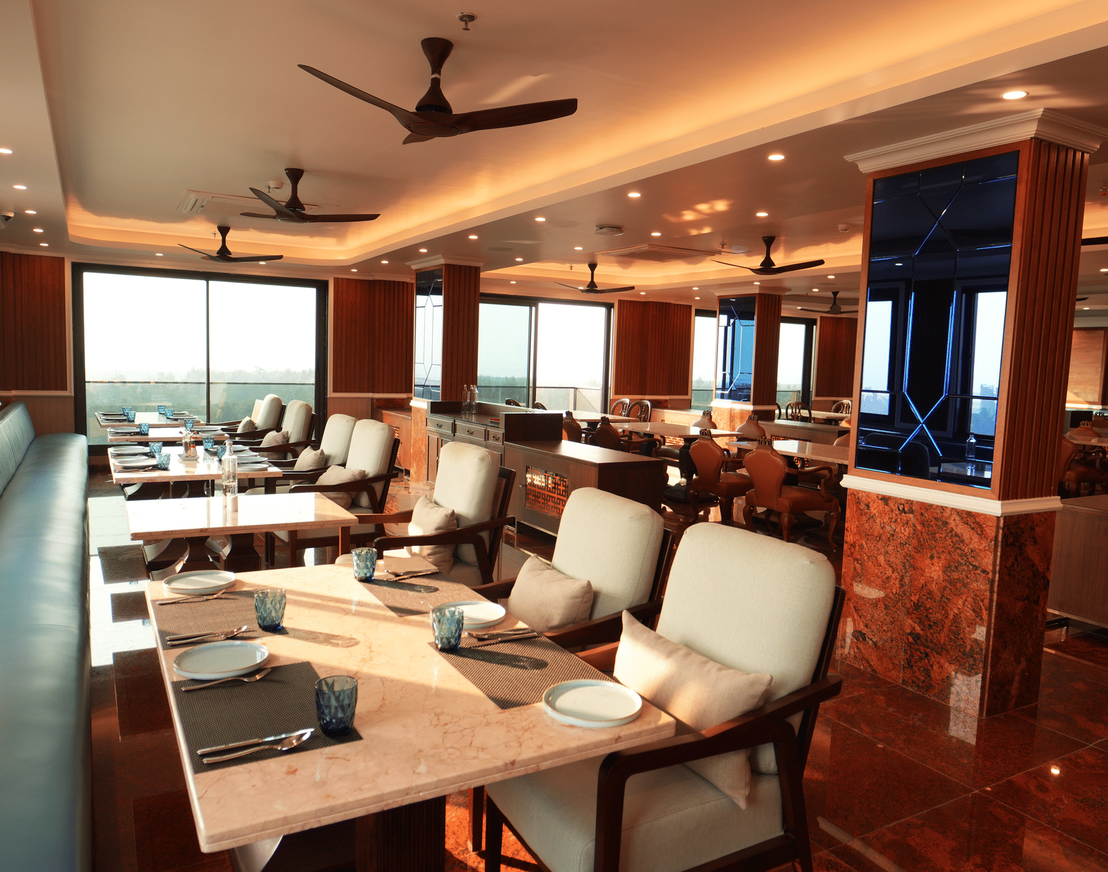 Indoor restaurant seating with cushioned chairs, tables set for dining, and ceiling fans overhead at MAYFAIR Bay Resort, Paradeep.