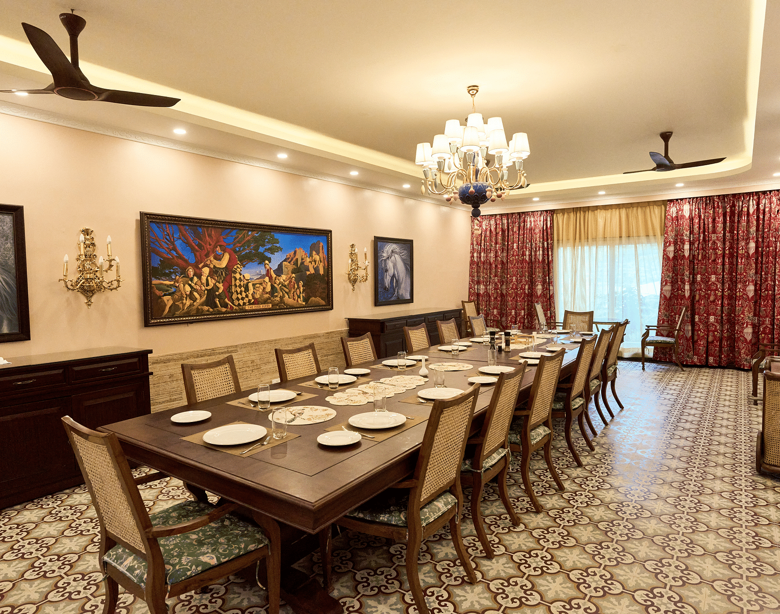 Diagonal view of a large dining table set for a culinary experience, with wall-mounted paintings, a chandelier, and large windows at Mayfair Manor, Alibag