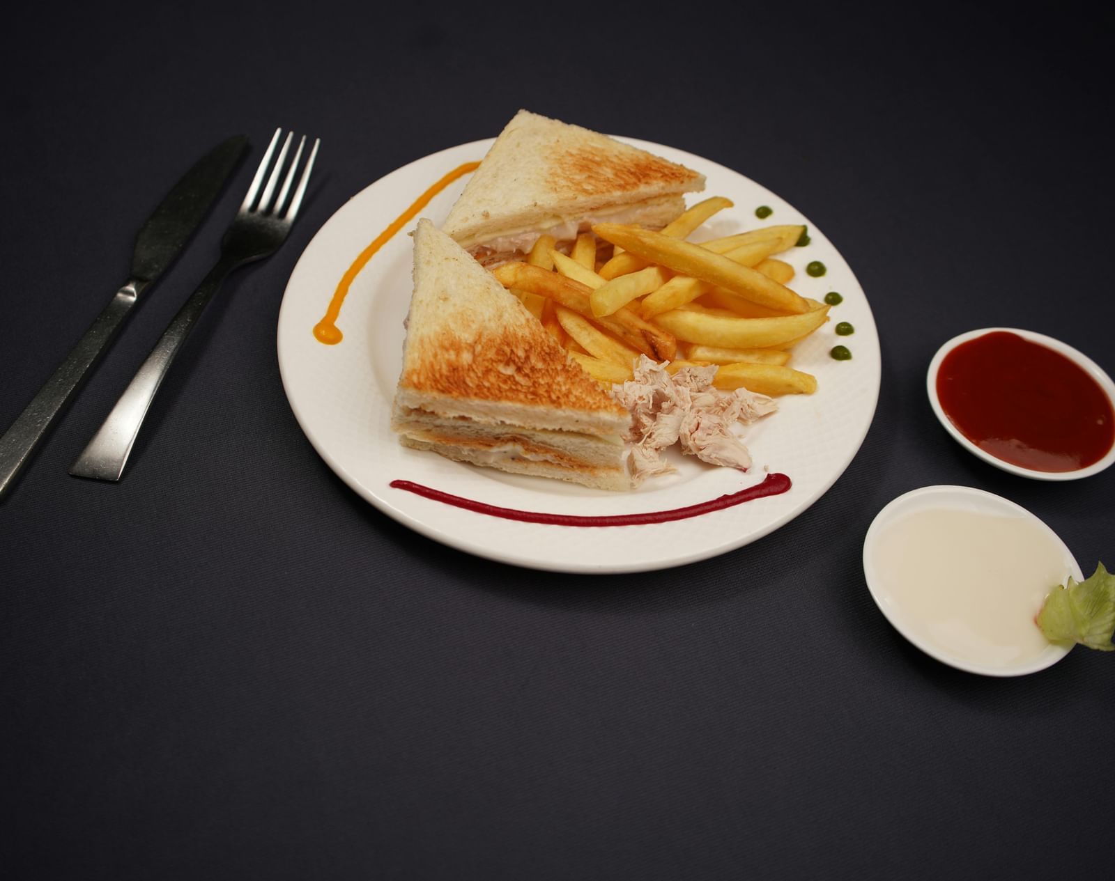 A plate of sandwich, is served with dipping sauces and a lime wedge on a dark surface.