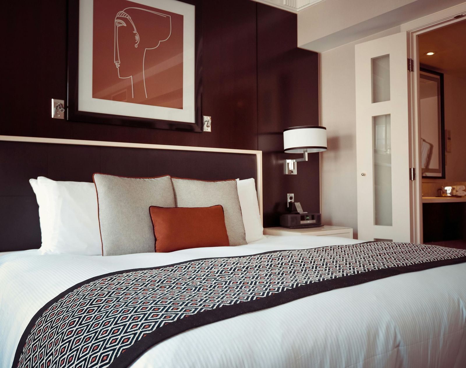 A hotel room displays a dark headboard, white linens, and a pop of colour with an orange pillow.