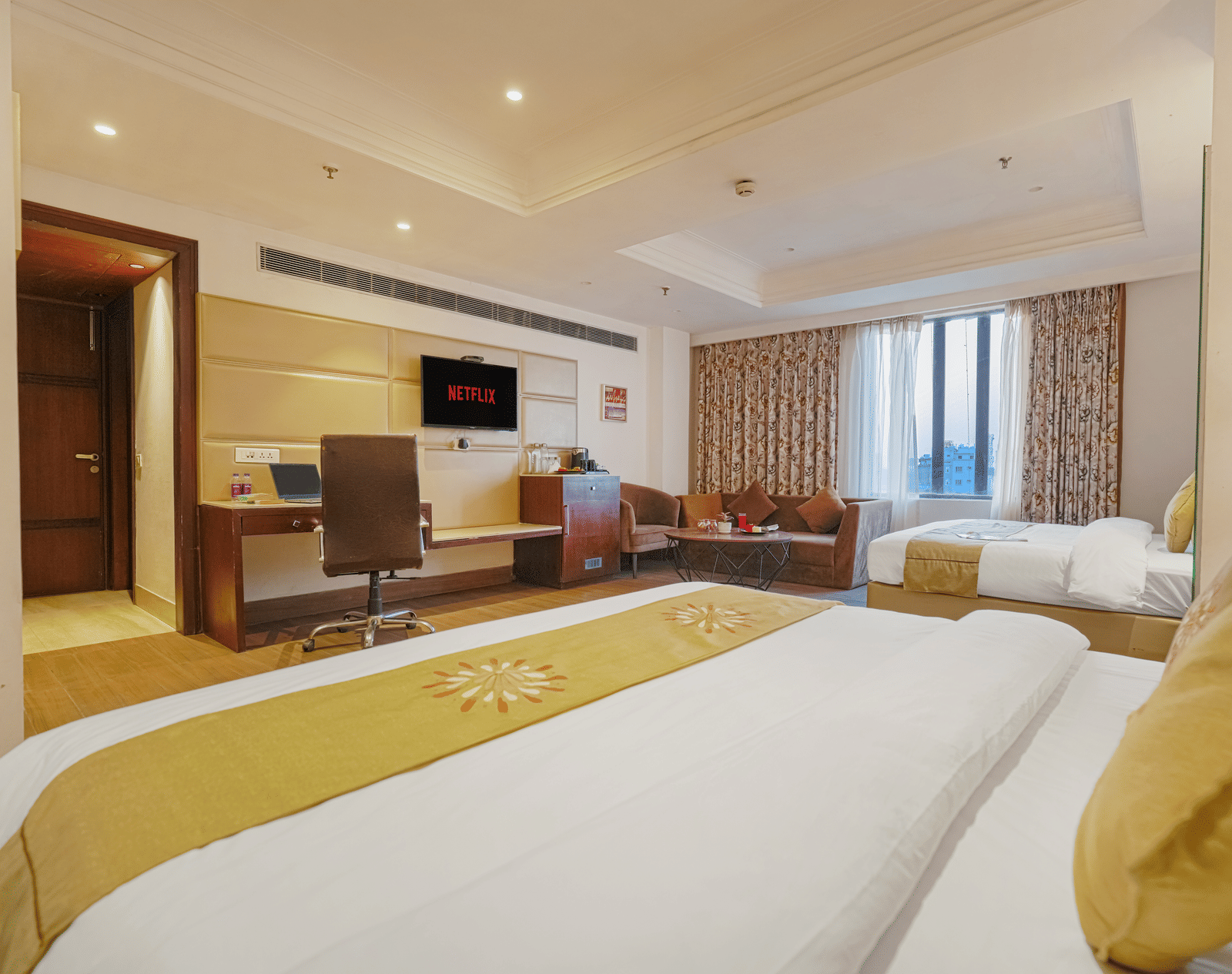 A spacious hotel room showing 2 beds with gold runners, a work desk, seating area, and wall-mounted television at BluSalzz Select City Centre, Amritsar.