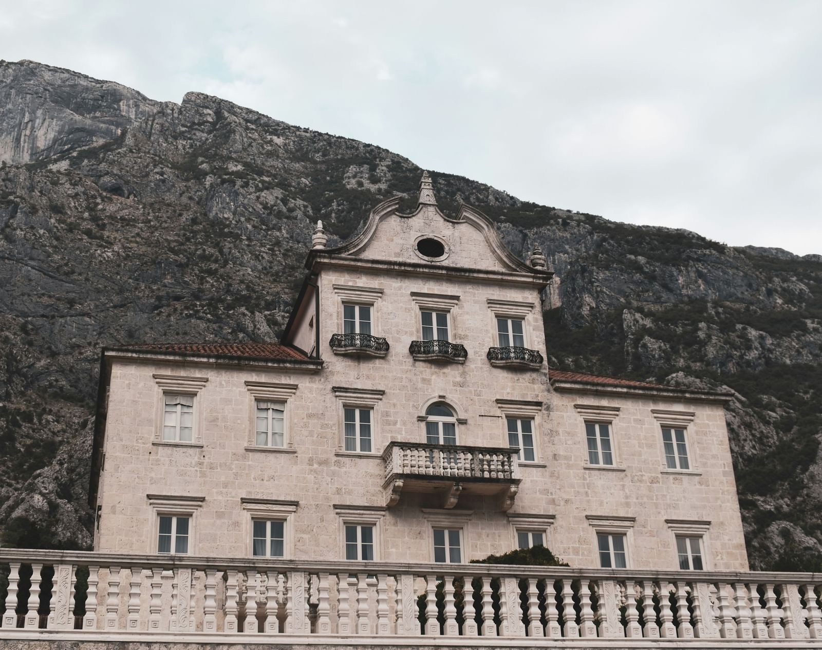 An old, majestic, multi-story building with a balcony and an arched entryway, set against a backdrop of a large mountain.