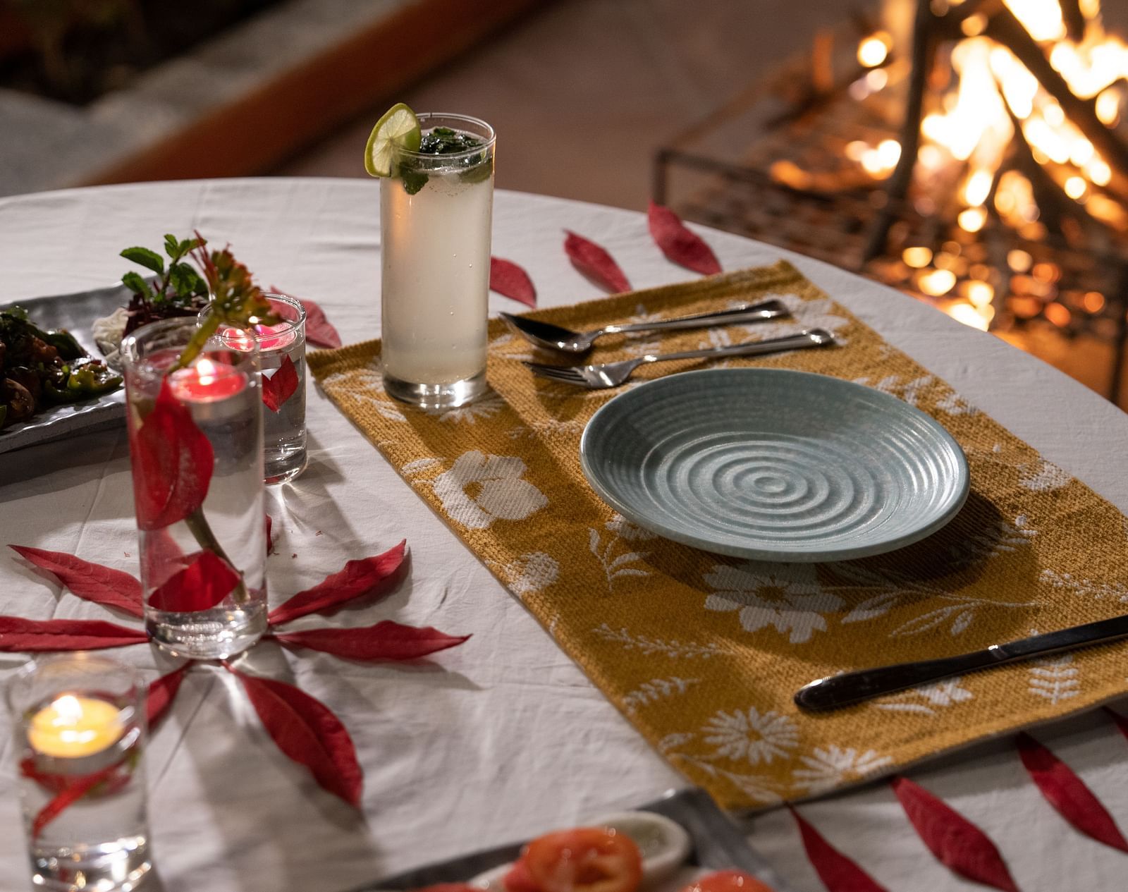 A close-up view of a table neatly set with food and cutlery, with a campfire in the background at BluSalzz Villas - The Green Roof, Bhimtal.
