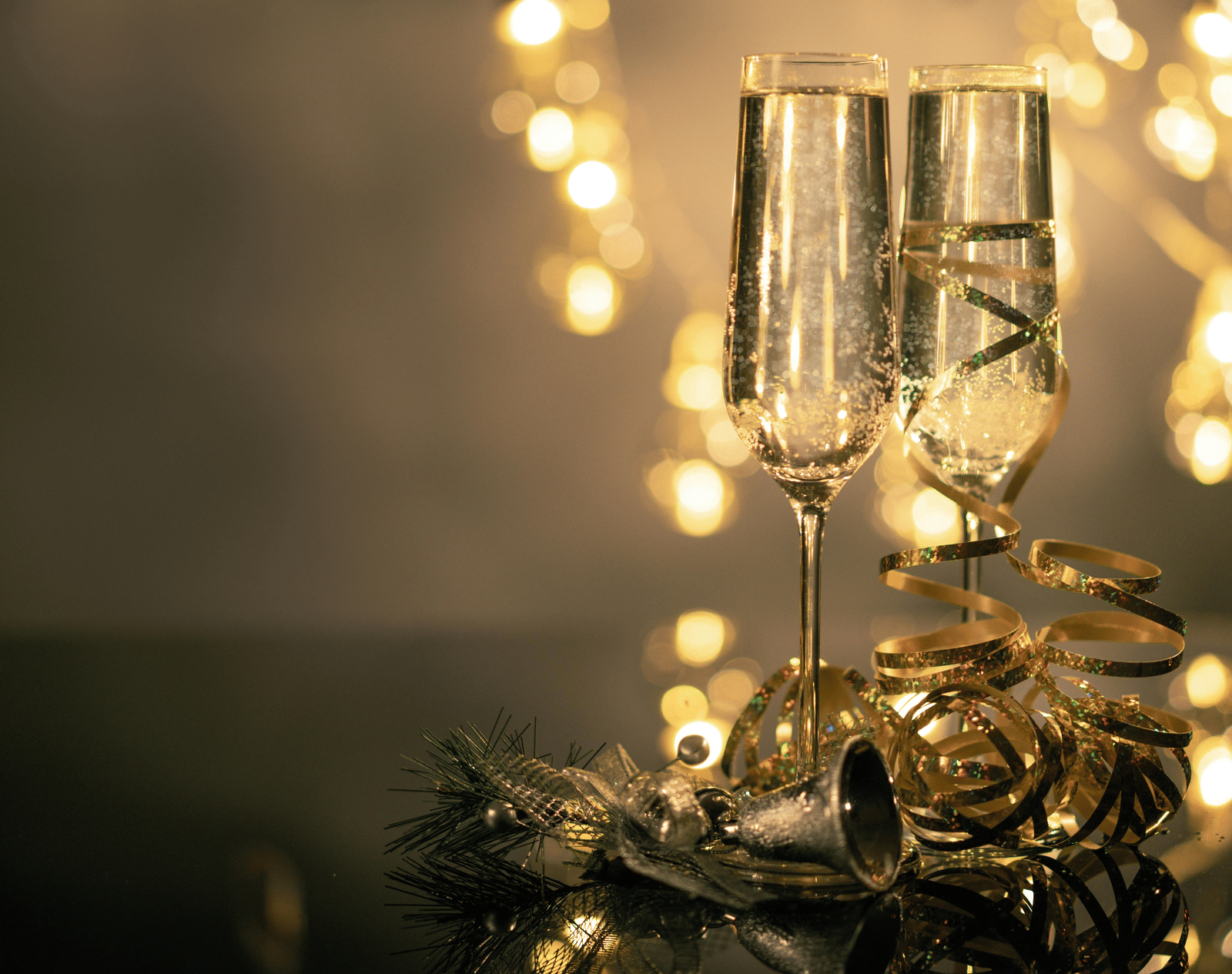 2 elegant champagne flutes standing on a table with gold tinsel, against a warm, blurred background of fairy lights.
