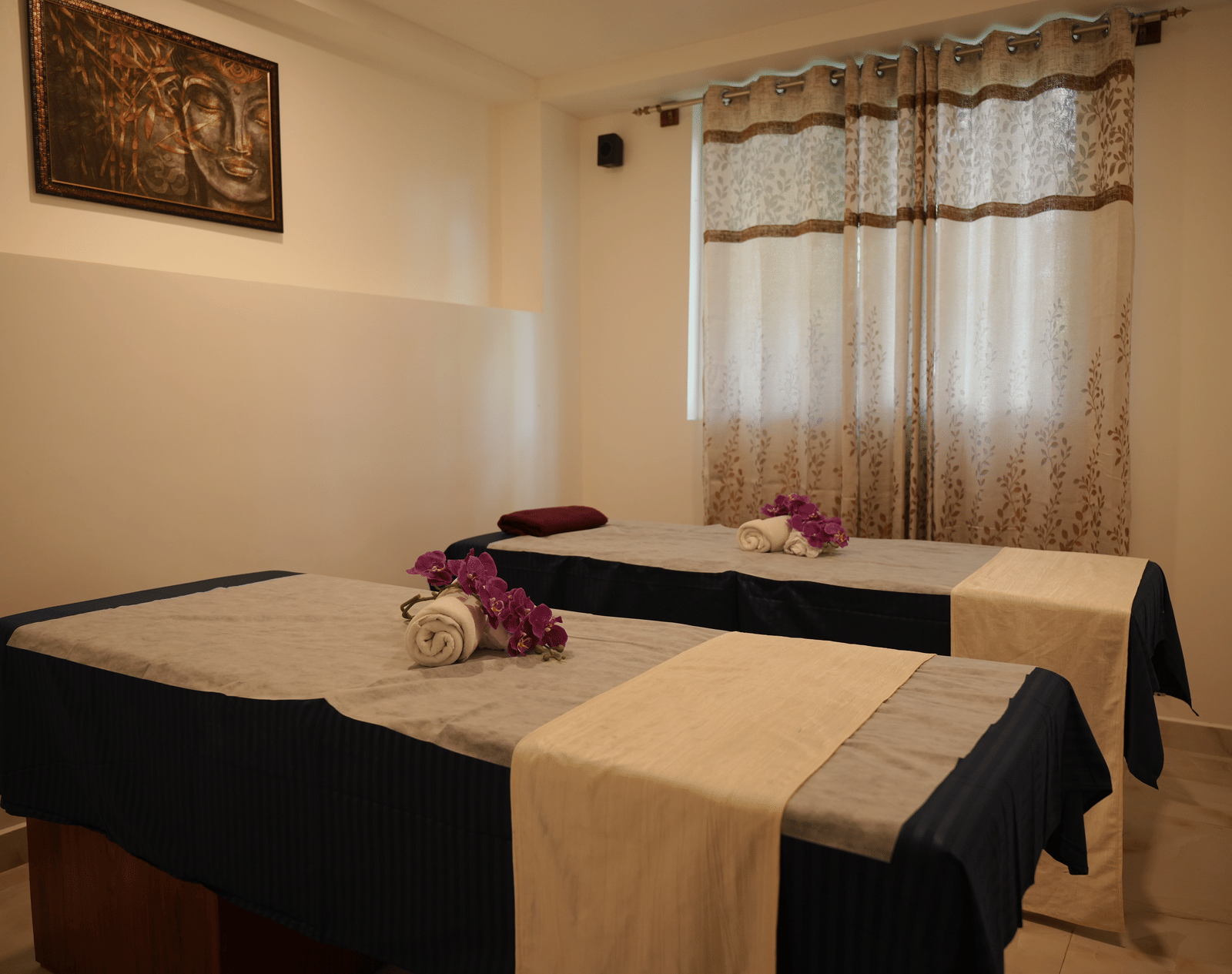 A spa treatment room containing 2 massage tables, sheer curtains, and simple wall decor at Coorg Orange Blossom Resort and Spa.