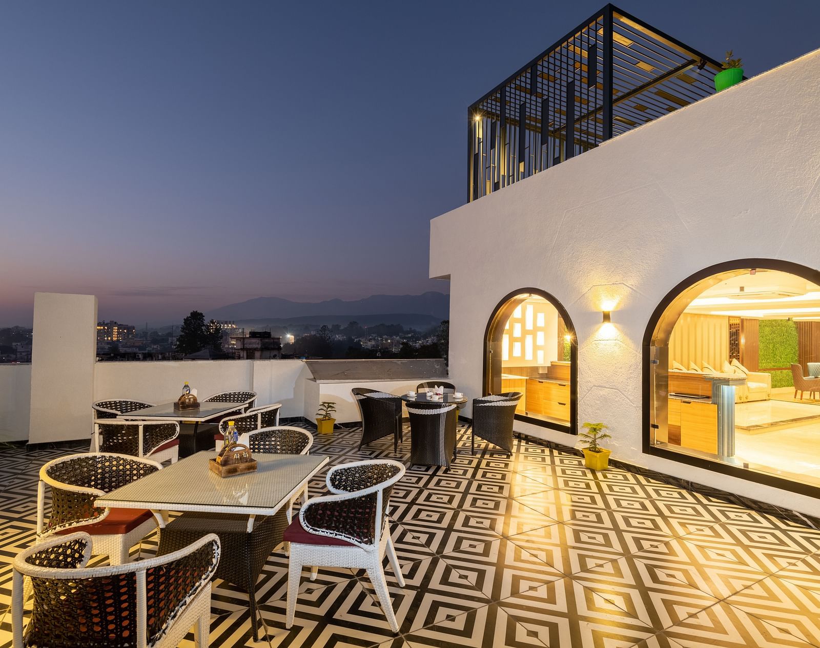Image of the rooftop restaurant at DLS Grand Luxotica, Dehradun during evening featuring chairs and tables arranged on a well-furnished floor