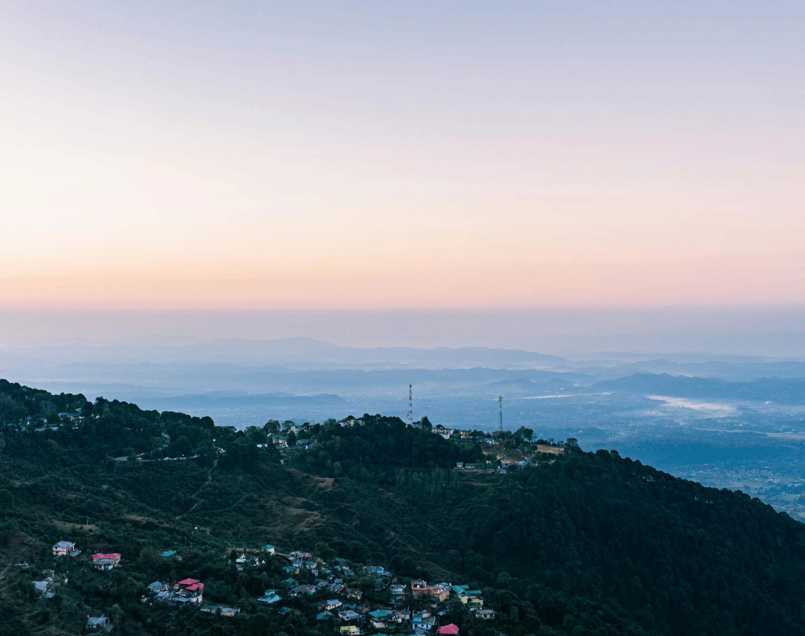 Scenic view of a hillside town with forested slopes, overlooking a vast valley and distant mountains under a pastel sky.