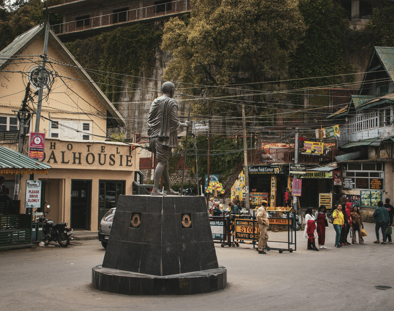 A statue of Gandhi in Dalhousie, set amidst bustling stalls and small eateries, with people walking through the lively market street.