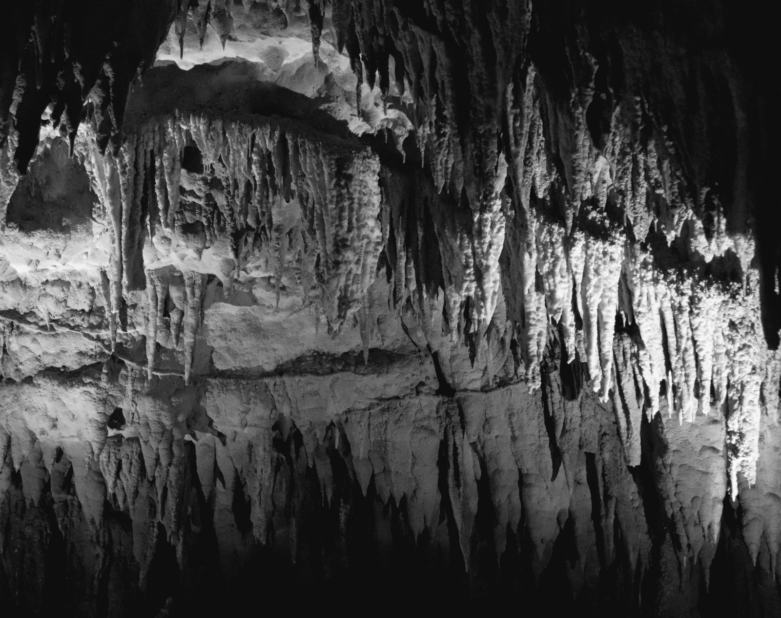 A black and white photograph of a cave with stalactites and their reflection in a pool of water.