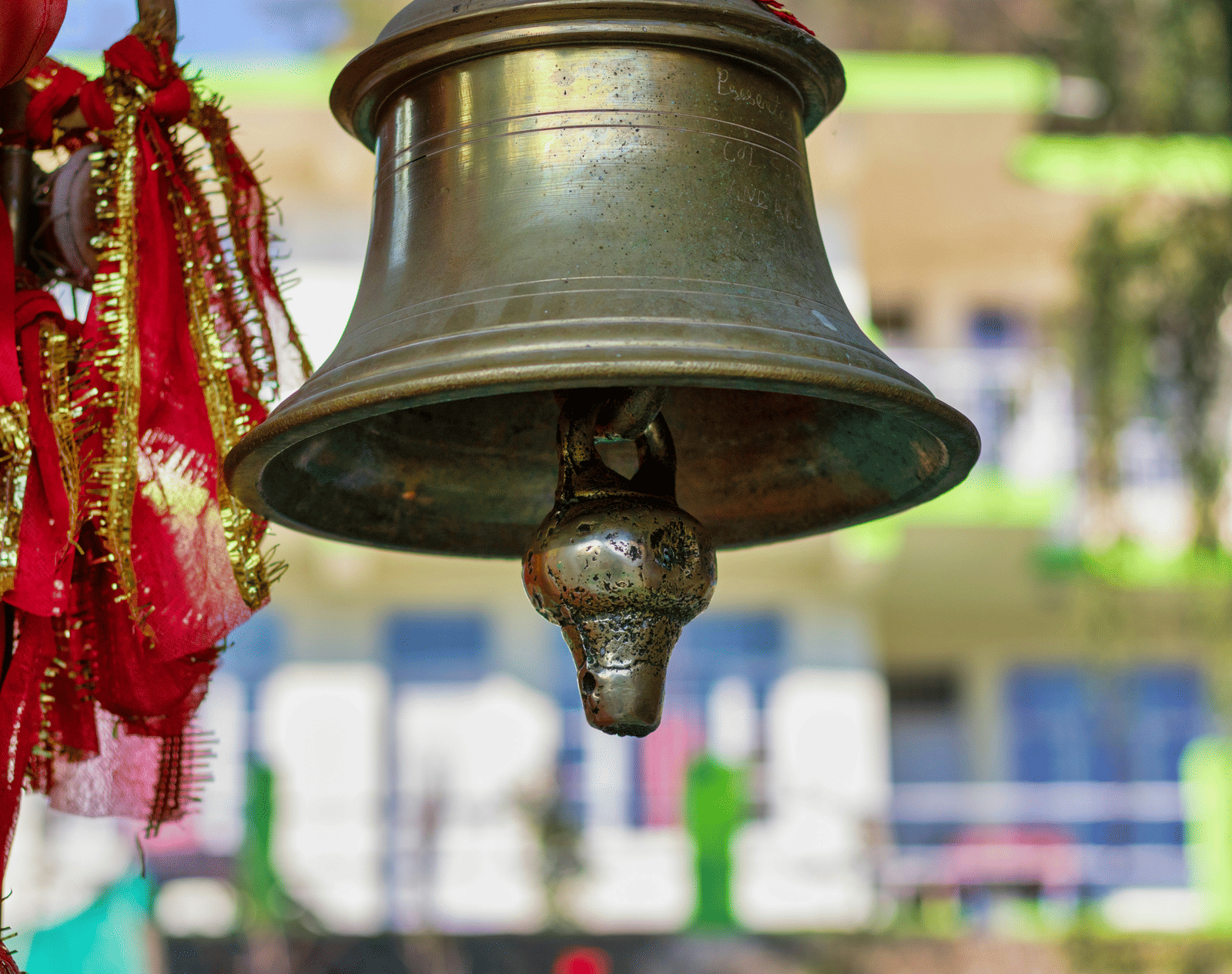 A temple bell decorated with red and gold ribbons hanging in the foreground with greenery behind it.