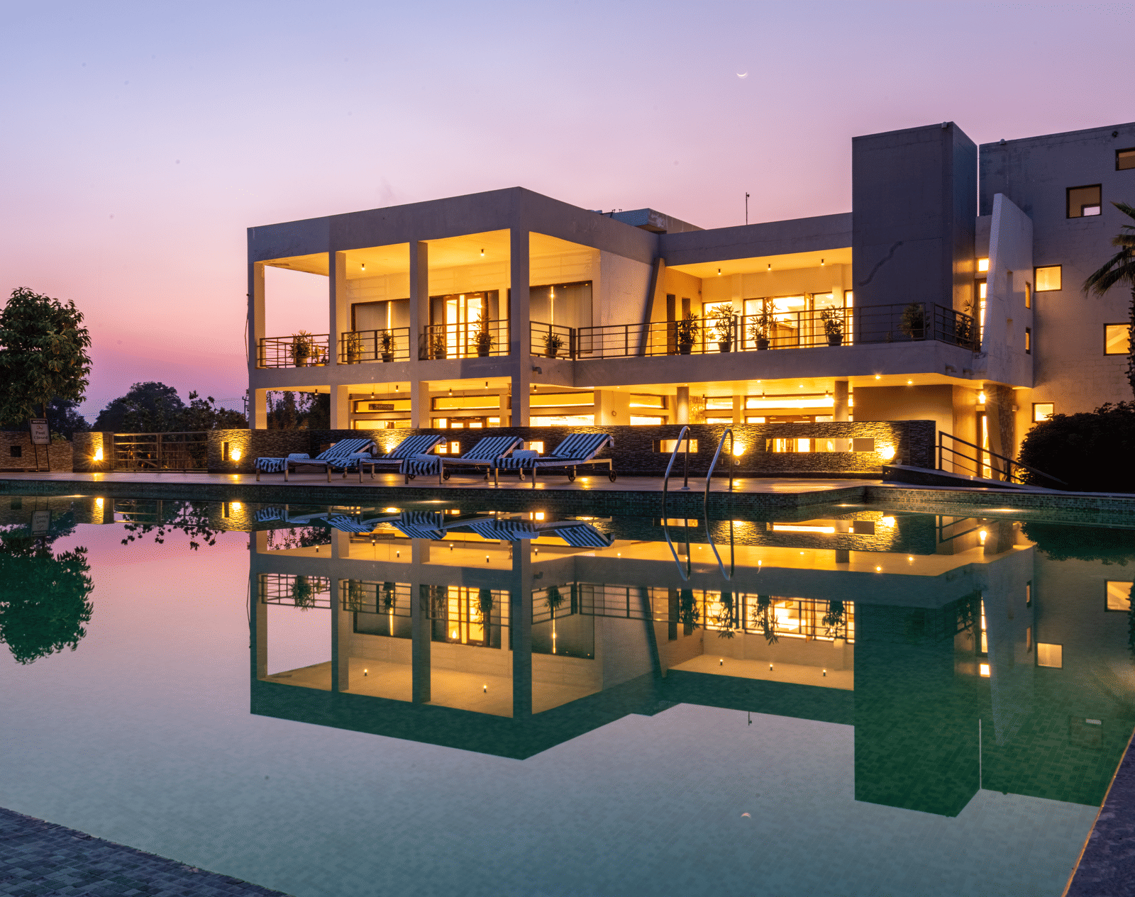 Facade view of DLS Forest Vines Resort, with warm light glowing from its windows and doors, reflected on the shimmering swimming pool beneath a clear sky.