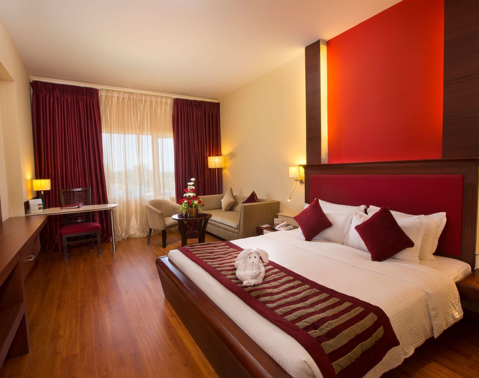 Bedroom with a rabbit made with towel on a large bed, table, chair and curtained window at  Hotel Atithi TGI Grand.