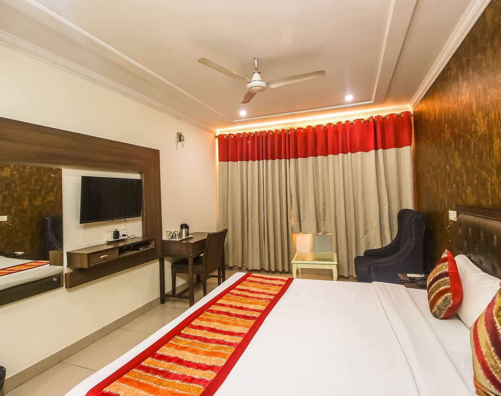 A hotel room with a plush bed featuring a patterned runner, a TV, a desk, a dark armchair, and a textured accent wall, lit by soft lighting. - Hotel Diamond Plaza, Chandigarh.