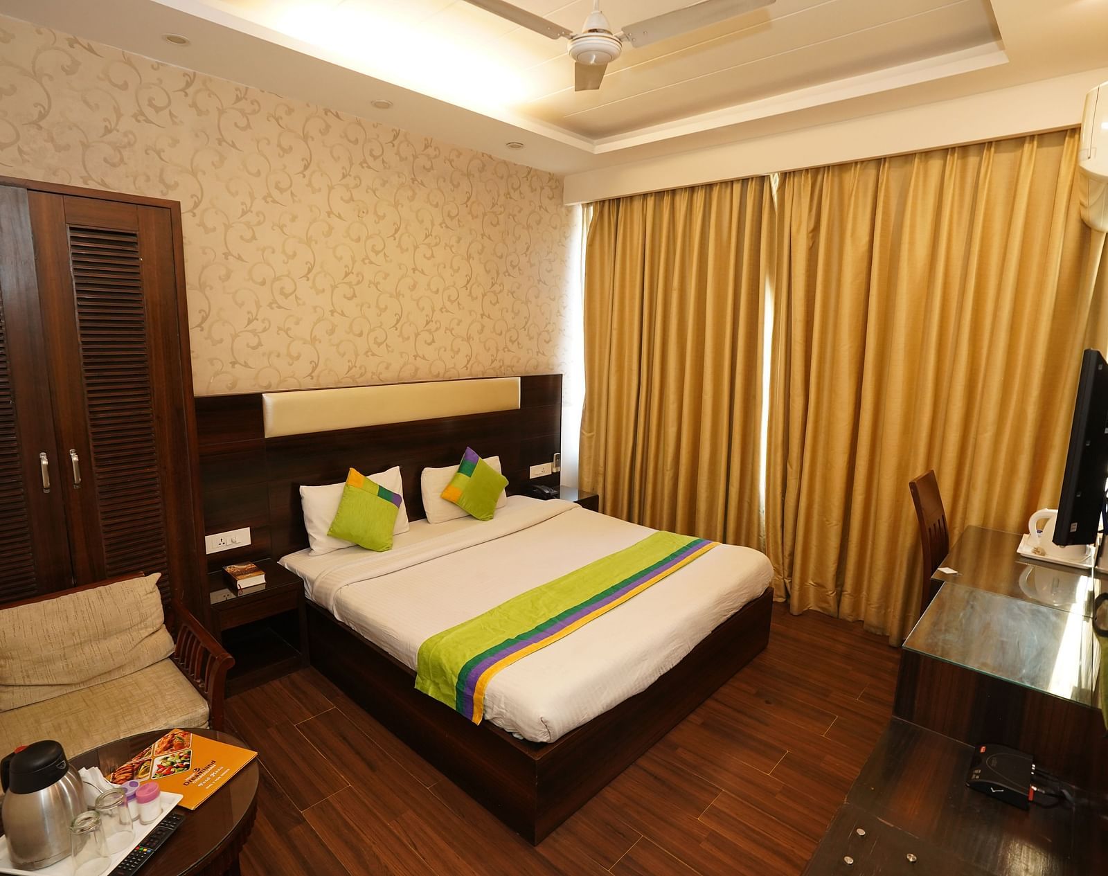 Deluxe Room featuring a king size bed with green accents, a wooden headboard, bedside tables, curtains, a desk, and a chair.- Hotel Dreamland, Chandigarh