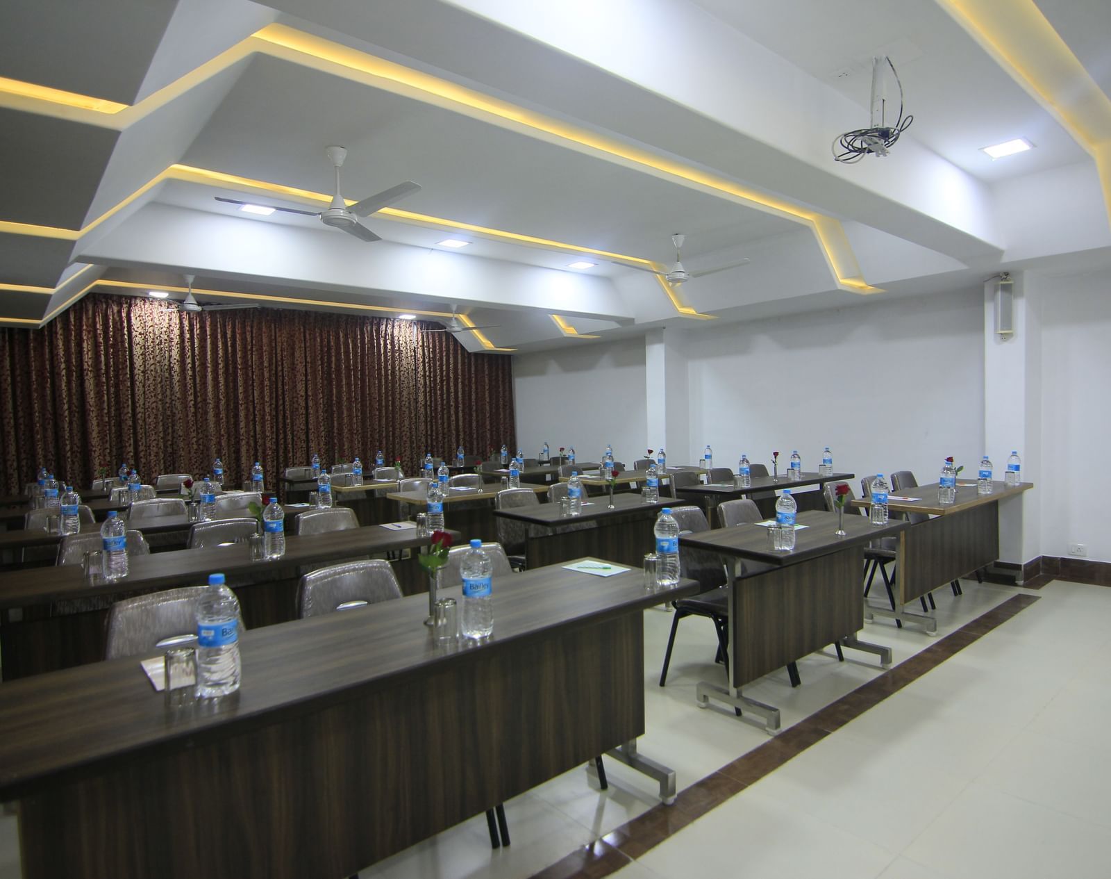 A well-lit conference room at Krushnai Resort with long rectangular tables arranged in class room style with chairs around them and a water bottle on each table.