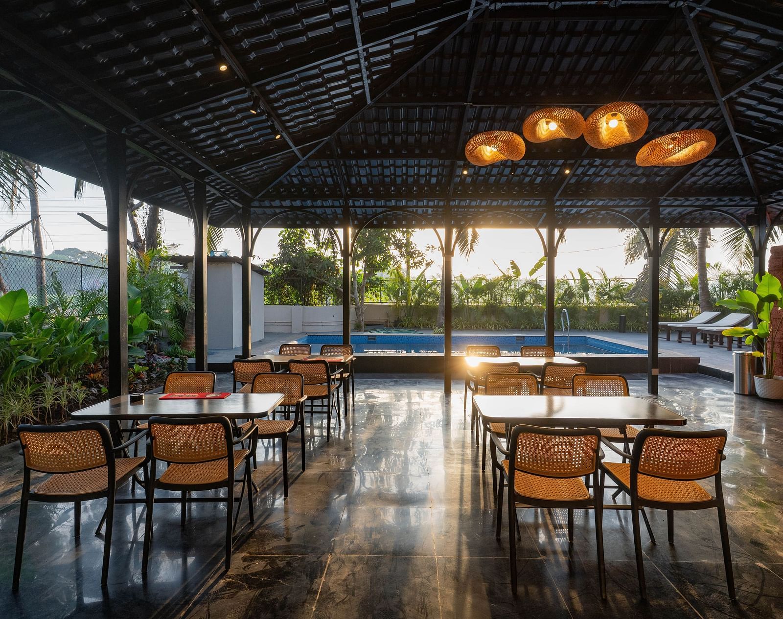 An outdoor dining area under a dark, peaked roof, open to the surrounding scenery. Several wooden tables and chairs are neatly arranged, with a glimpse of a swimming pool visible in the background under the bright sky at Kssemya