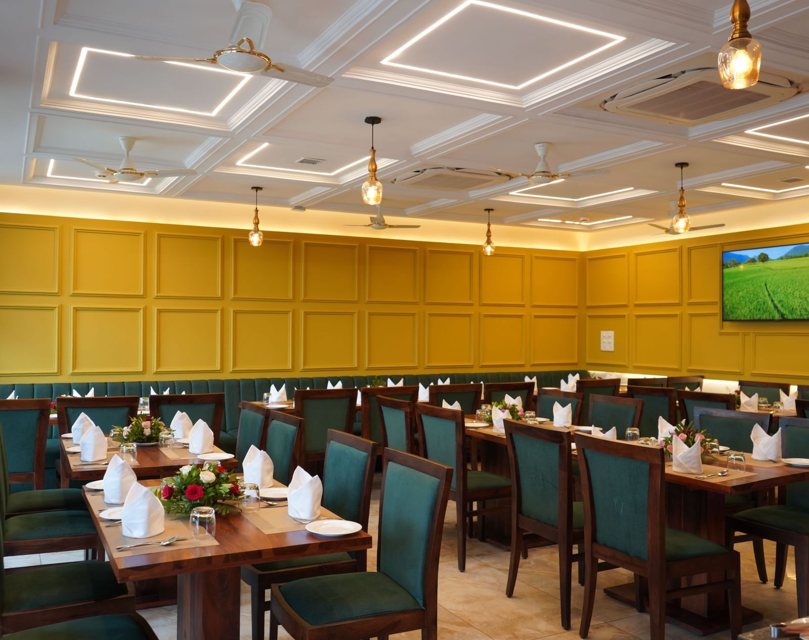 A wide shot of a dining hall with multiple tables set for a meal. The chairs are dark green, and the walls are yellow. A TV is visible on the far wall.