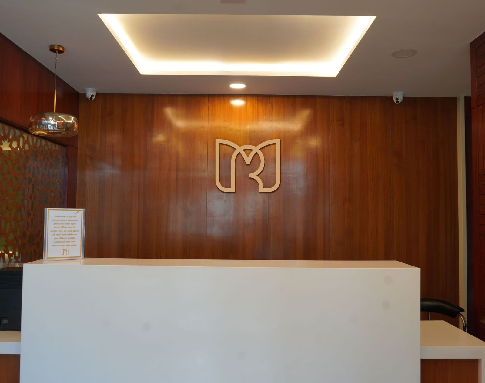 Reception area of Mertiya Residency with a minimalist white counter, warm wooden wall panels, and a stylish illuminated logo in the background.