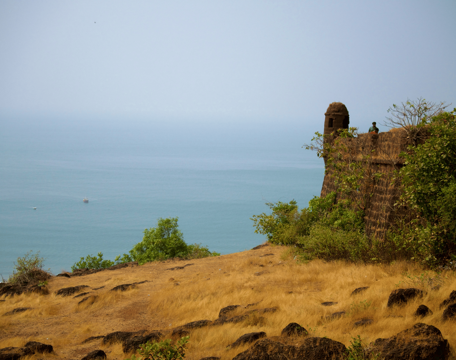 The ruins of a stone fort perched on a dry, grassy cliff, overlooking the calm blue sea under a bright sky.
