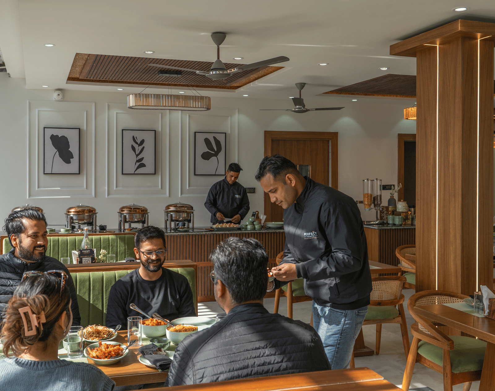 4 people sitting with 2 office staff, one serving, and another standing by the reception, at a dining table inside Perfectstayz Premium Kapilaz, Bhimtal.